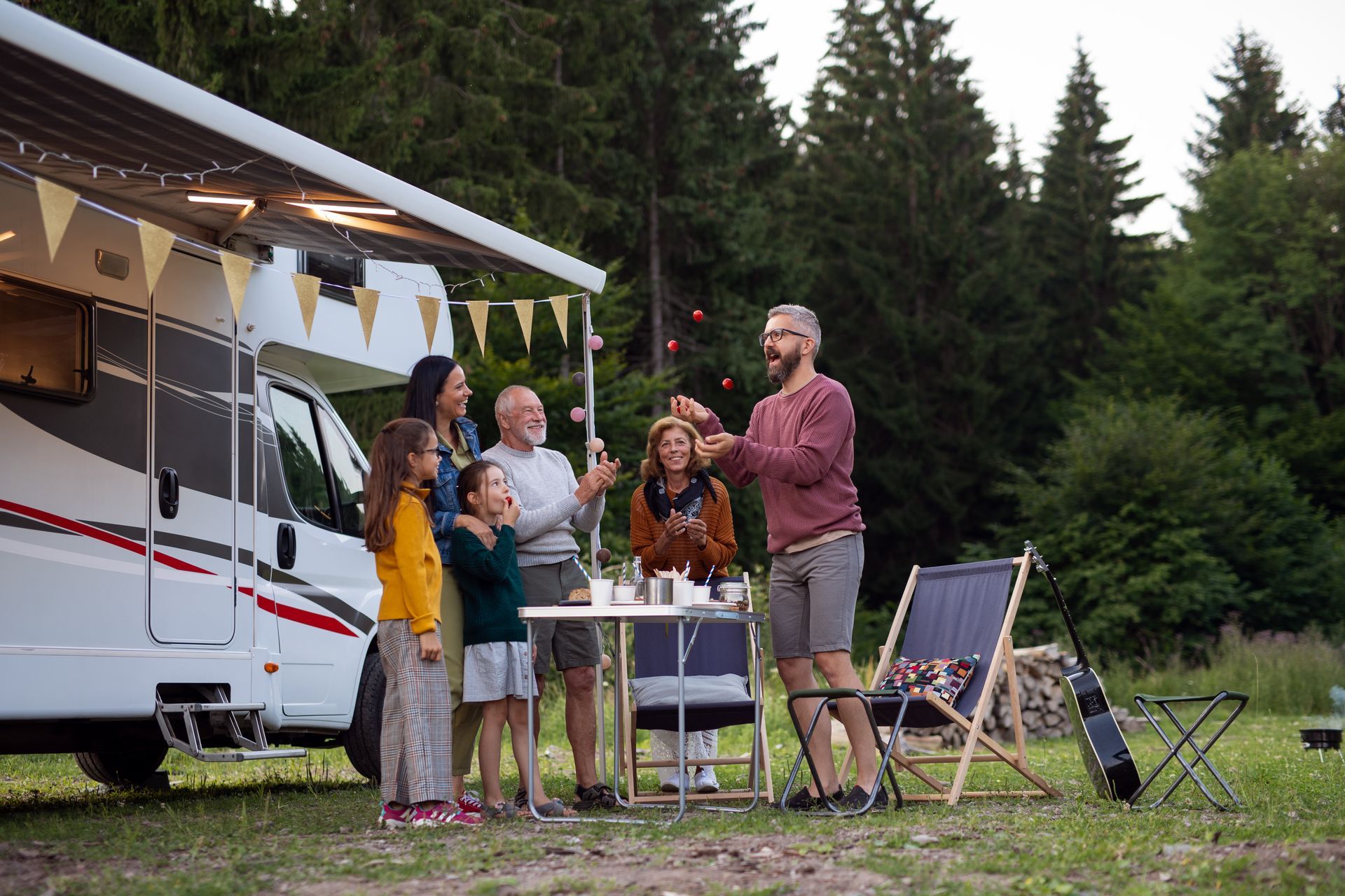 People chatting and holding drinks beside a camper at a forest campsite at dusk