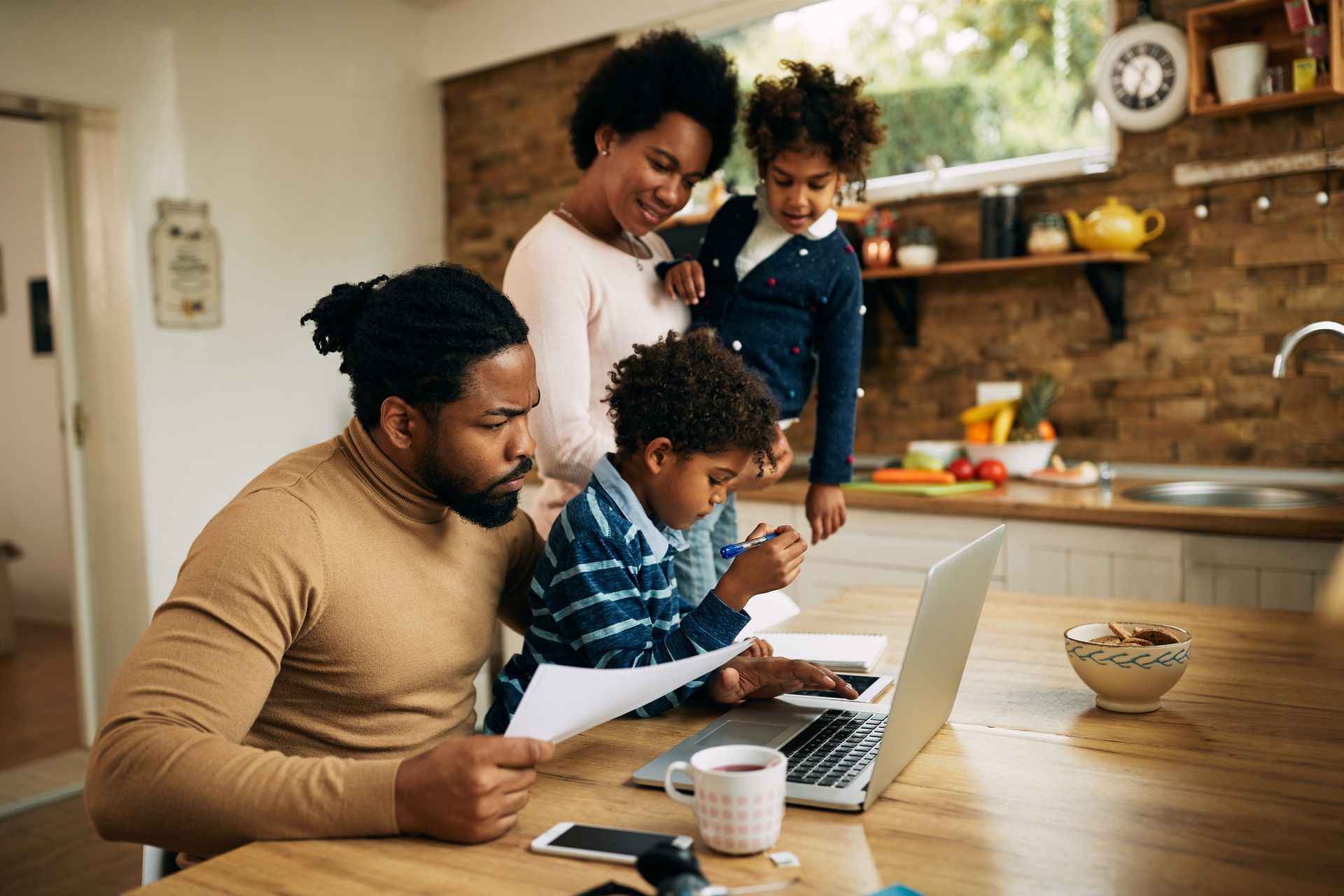 Family gathered at a kitchen table, using a laptop and papers while a child stands nearby.