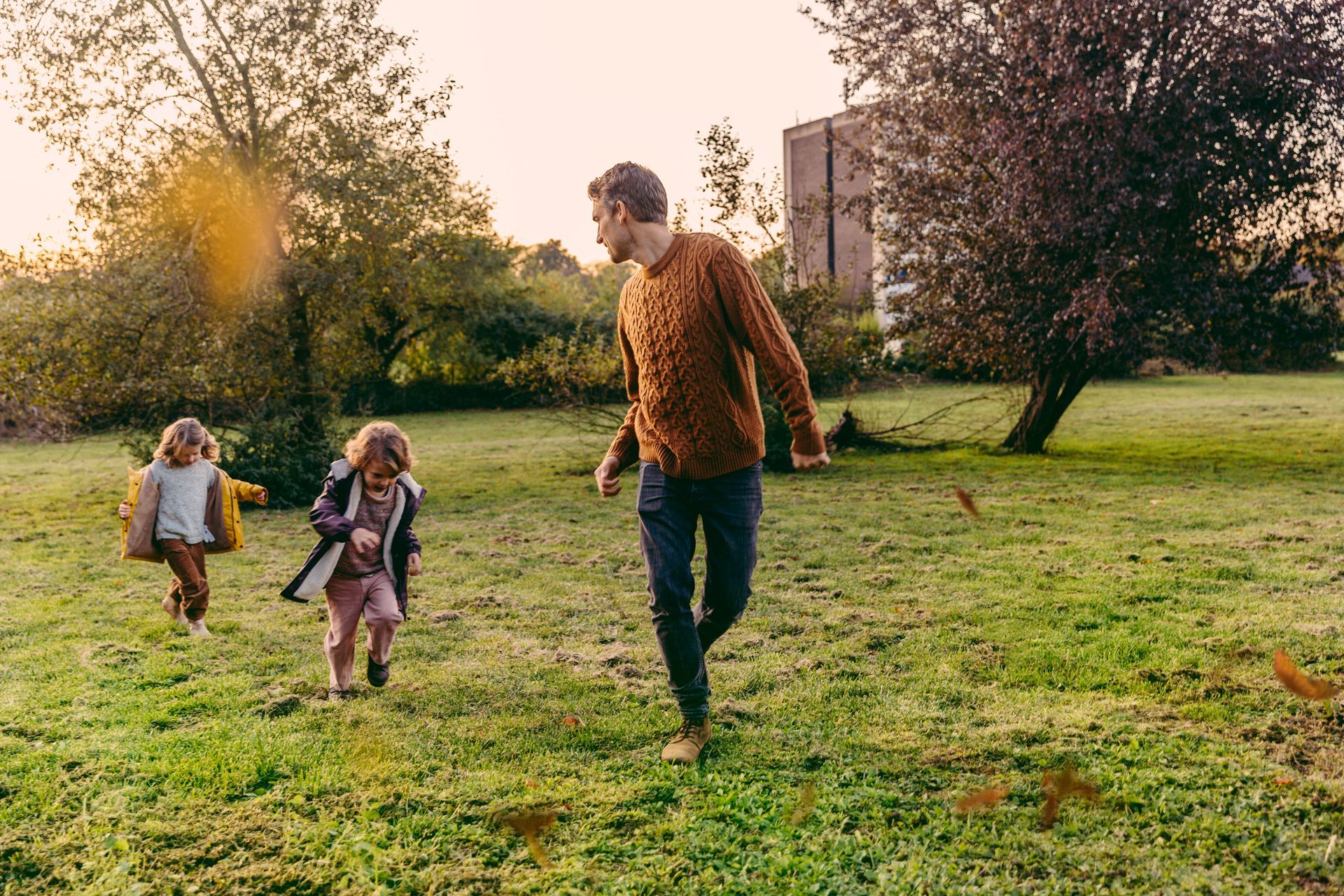 Two children and an adult run across a grassy field at sunset near trees and a building