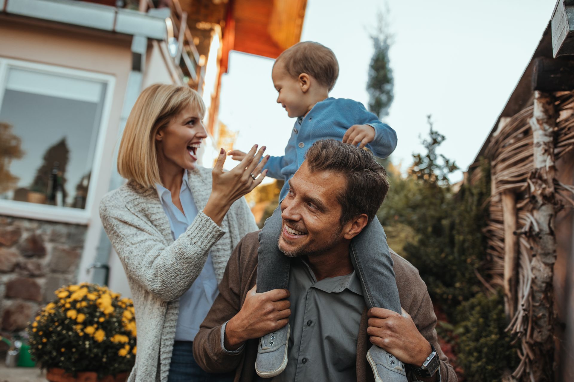 Family smiling outside a home, woman clapping while a child rides on a man’s shoulders