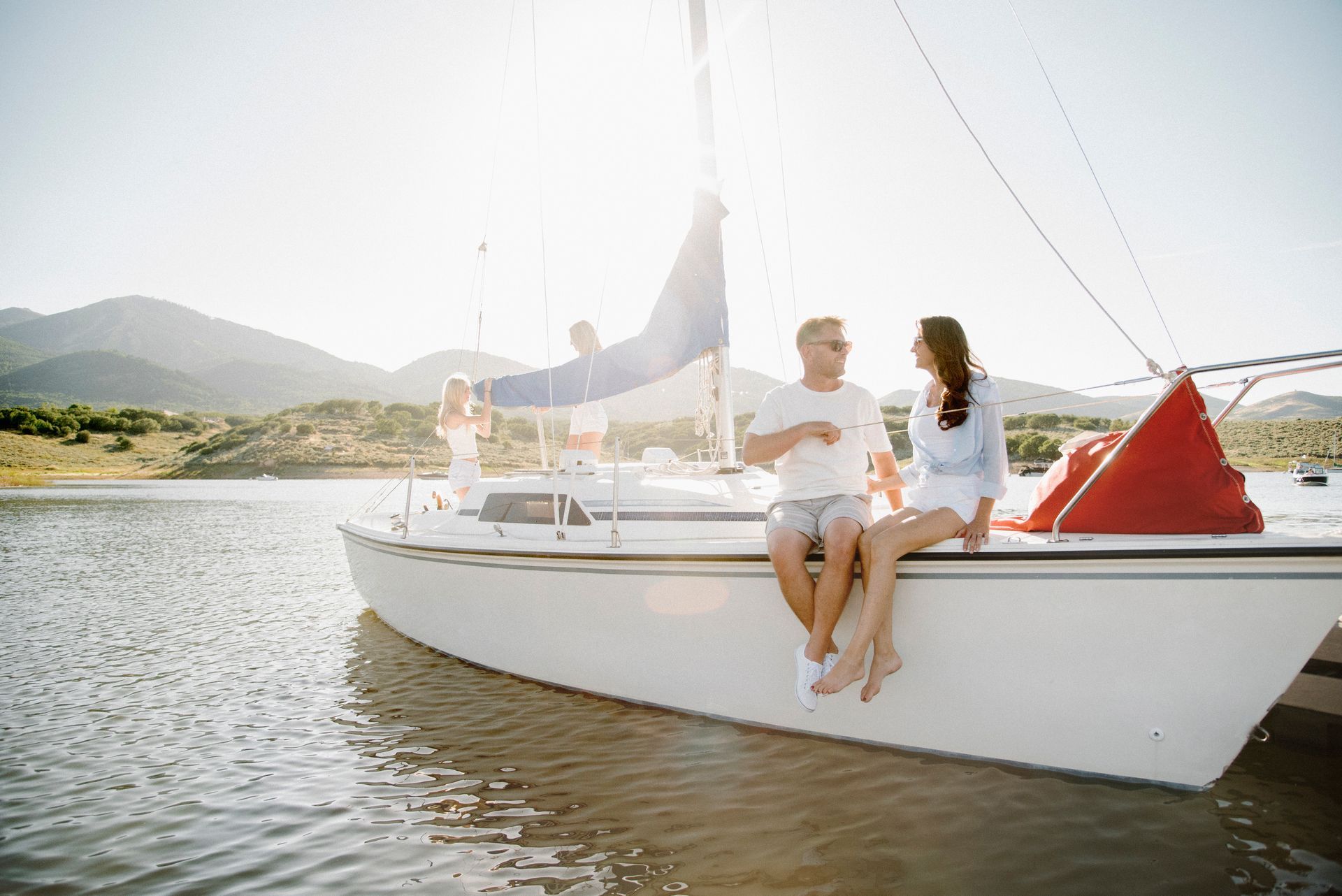 Two people sitting on a white sailboat in calm water with mountains in the background