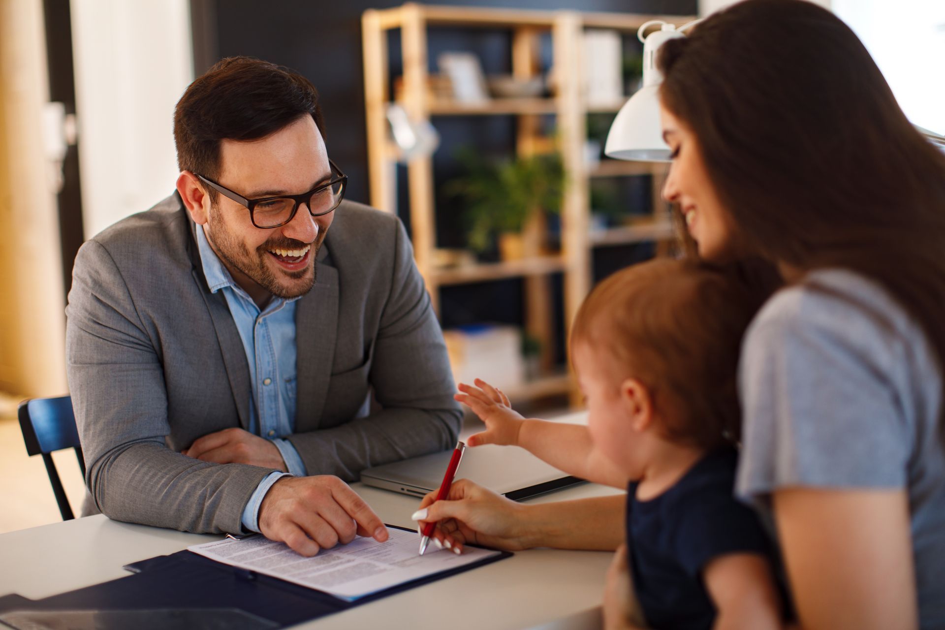 Two people and a child talking with a man at a desk in a bright office, with papers and a pen on the table.
