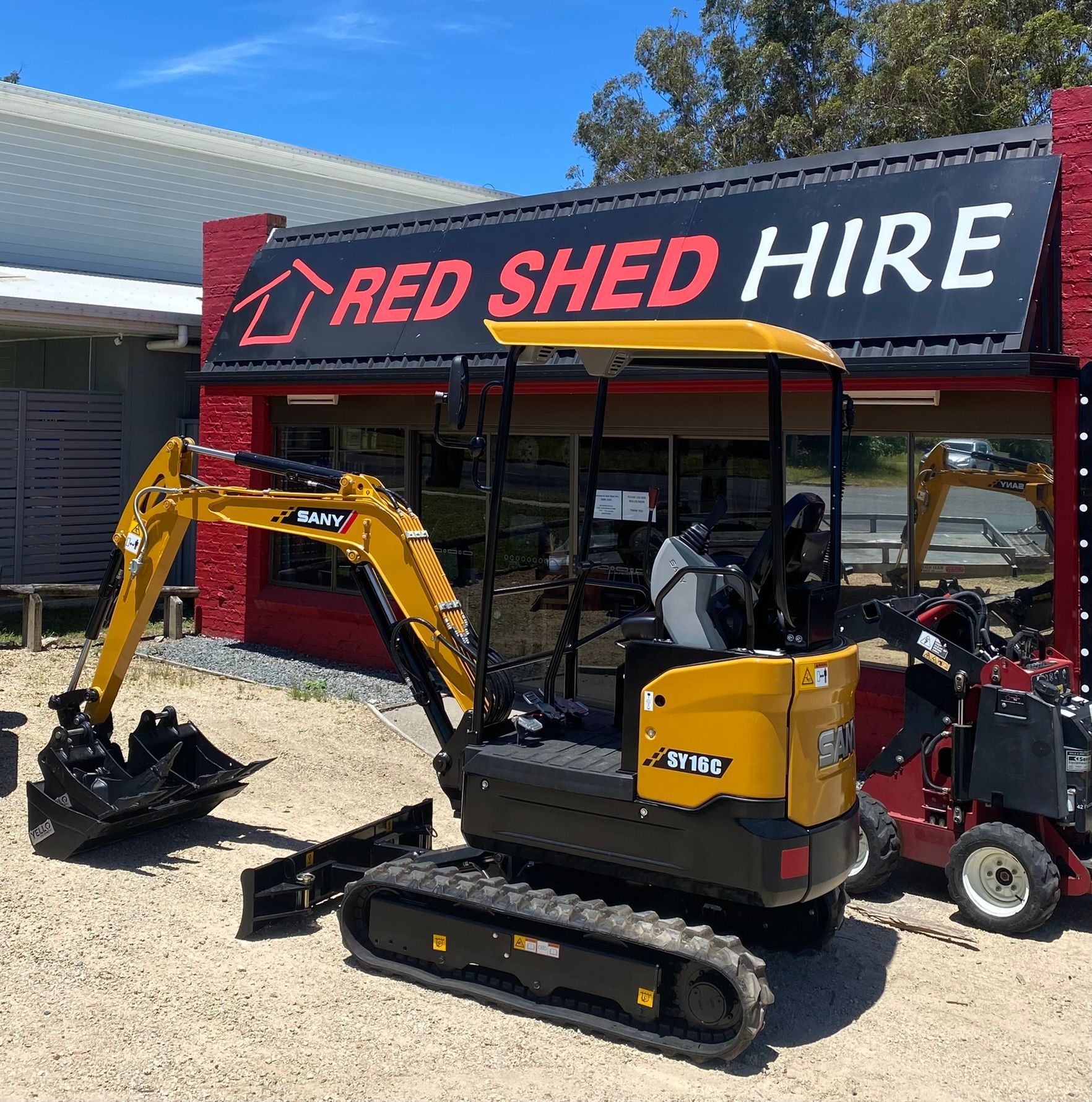A Yellow Excavator Is Parked In Front Of A Red Shed Hire Store — Red Shed Hire In Wauchope, NSW
