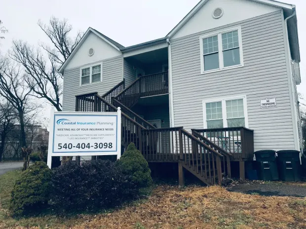 A two-story light gray building with a wooden staircase and a white business sign in the front yard.