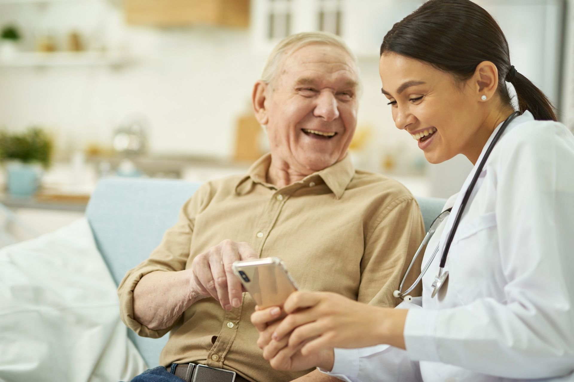 A healthcare professional in a white coat sits with a smiling person, both looking at a smartphone together indoors.