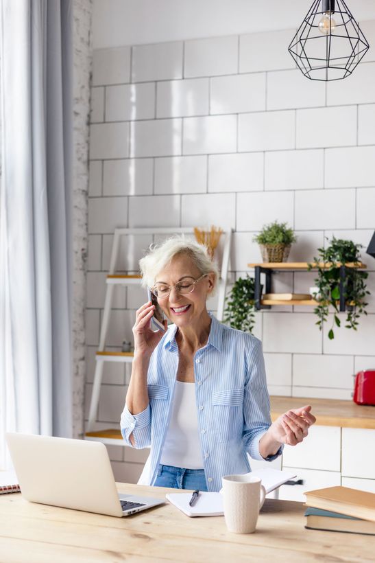 A person in a striped shirt sits at a desk with a laptop and phone, smiling while working in a bright kitchen.