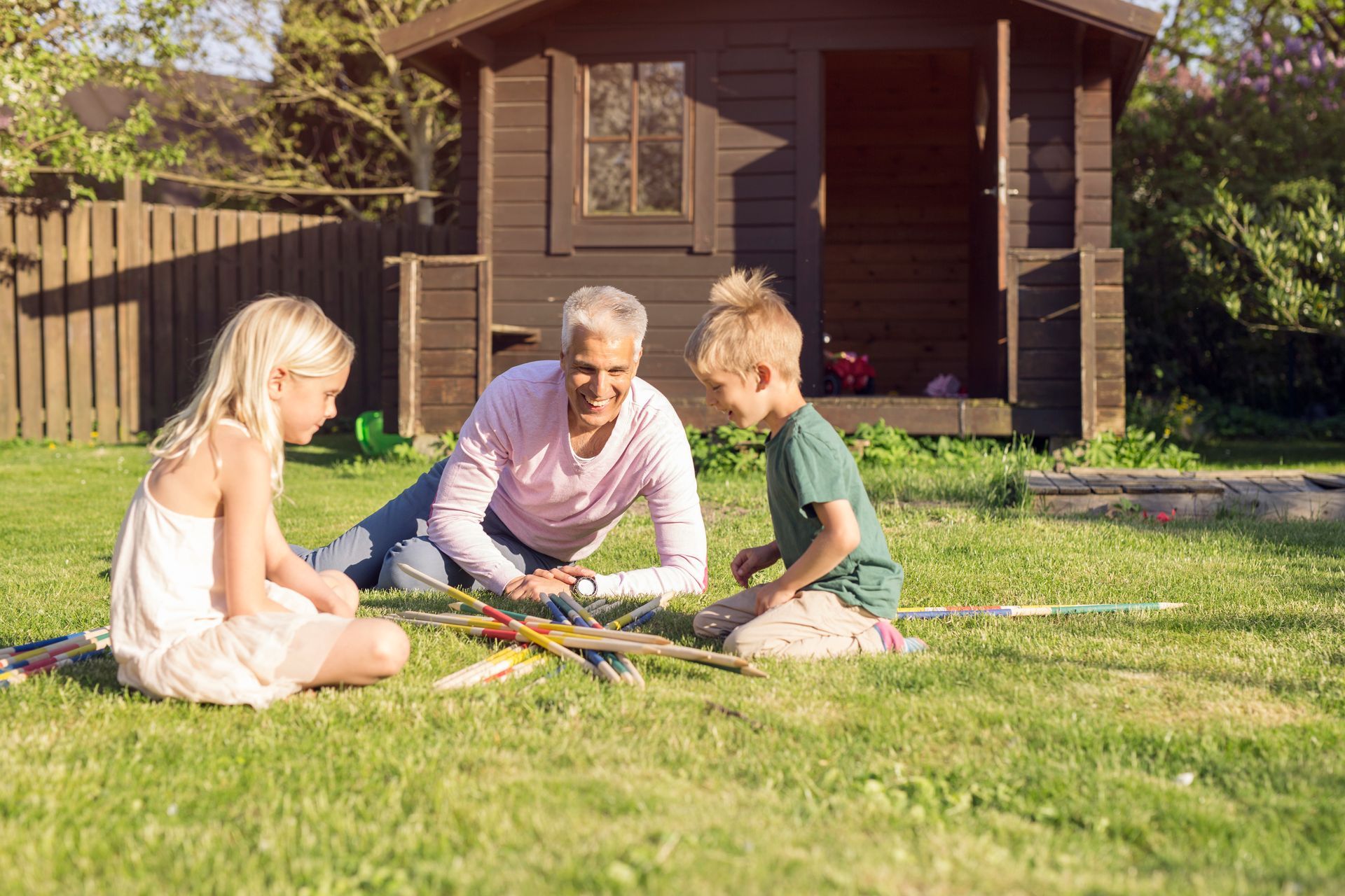 An adult and two children play with sticks on the grass in front of a wooden garden shed on a sunny day.