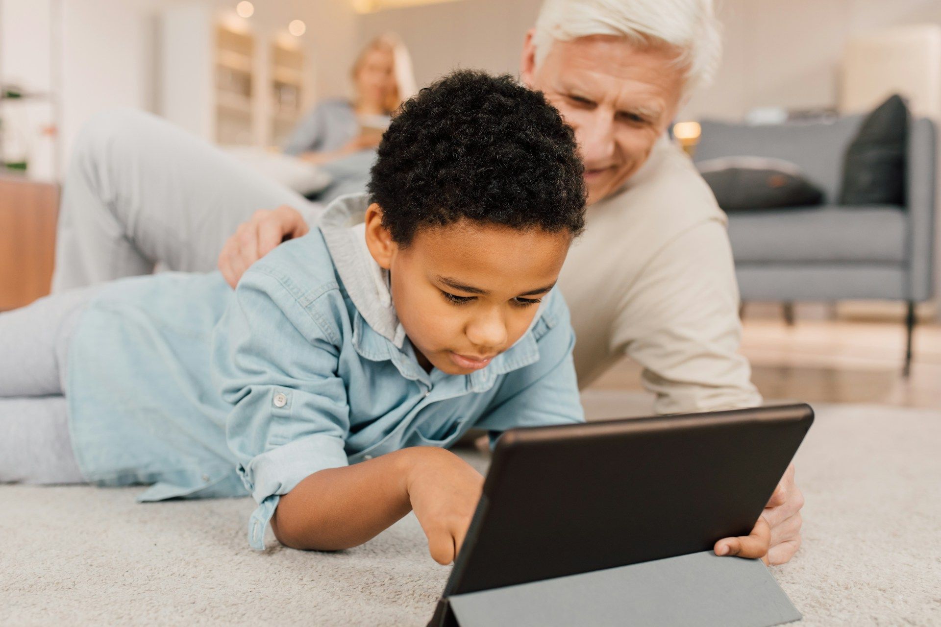A person and a child lying on a carpeted floor, focusing together on a tablet screen.