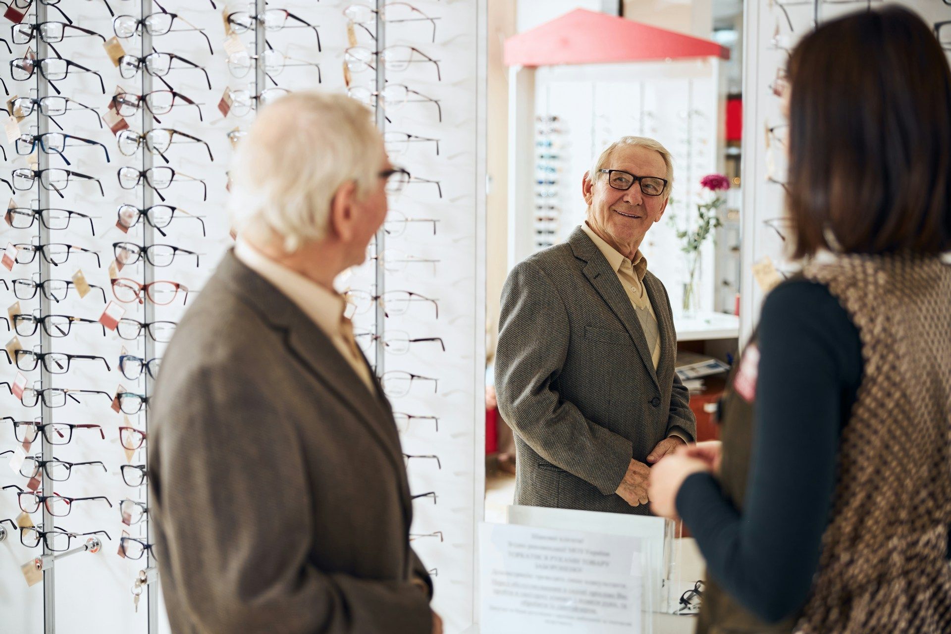 A person tries on glasses while looking in a mirror at an optical store, assisted by an employee.