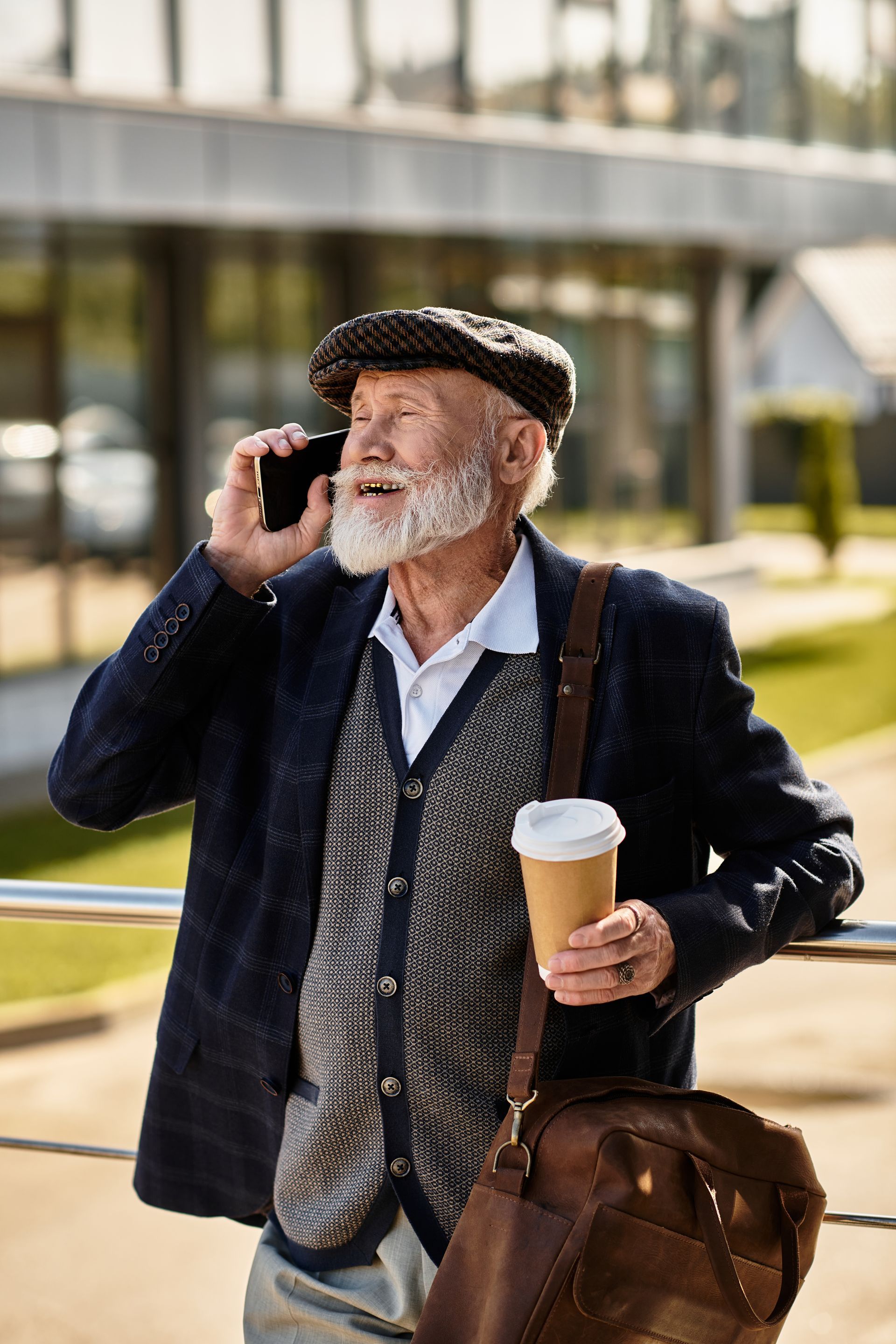 A person in a flat cap and blazer talking on a phone, holding a paper coffee cup while leaning on a metal railing.