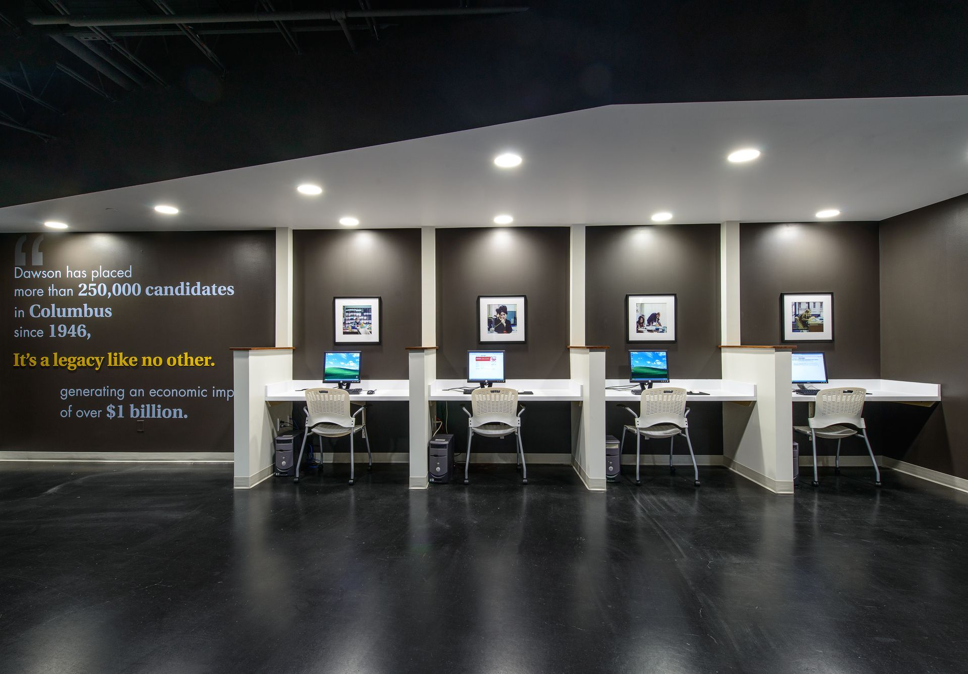 A row of four computer workstations in a dark-walled office, each with a chair, monitor, and framed artwork above.