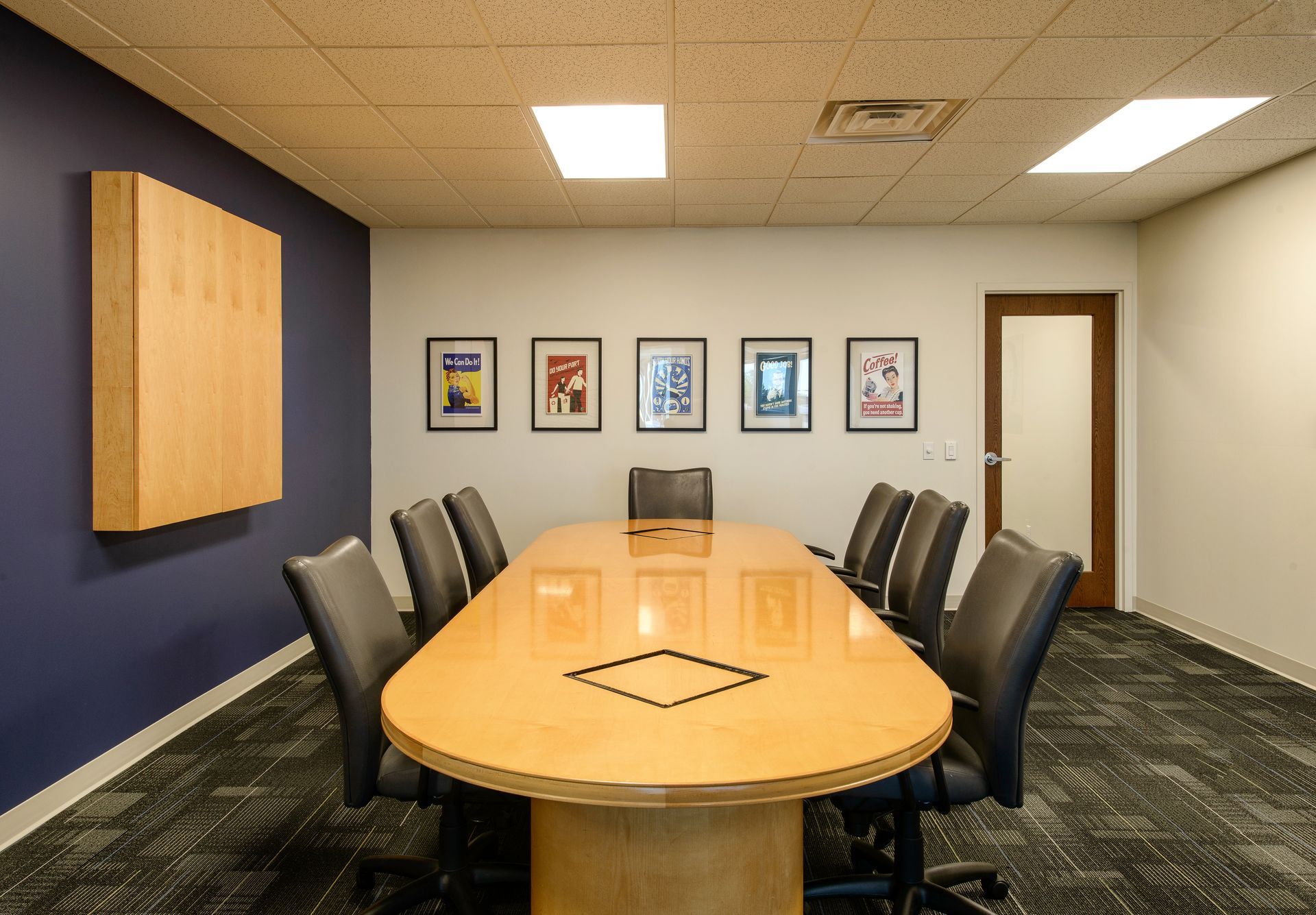 A conference room with a long light-wood table, ten office chairs, and five framed posters on a white back wall.