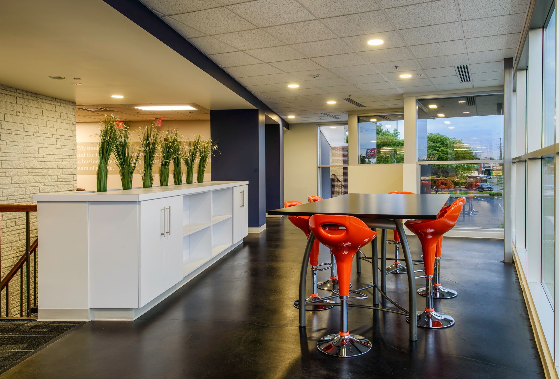 A modern office lounge with a long white counter, indoor plants, and a high table with orange stools on a dark floor.