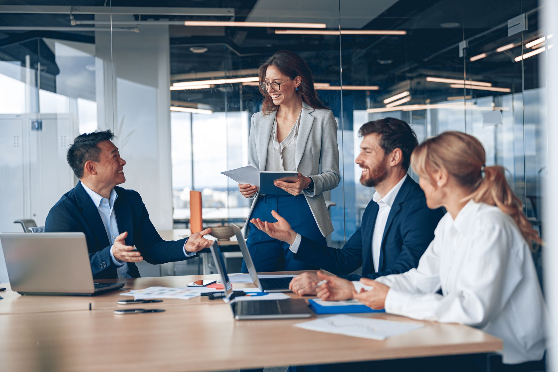 Four colleagues in business attire collaborating during a meeting in a modern, glass-walled office.