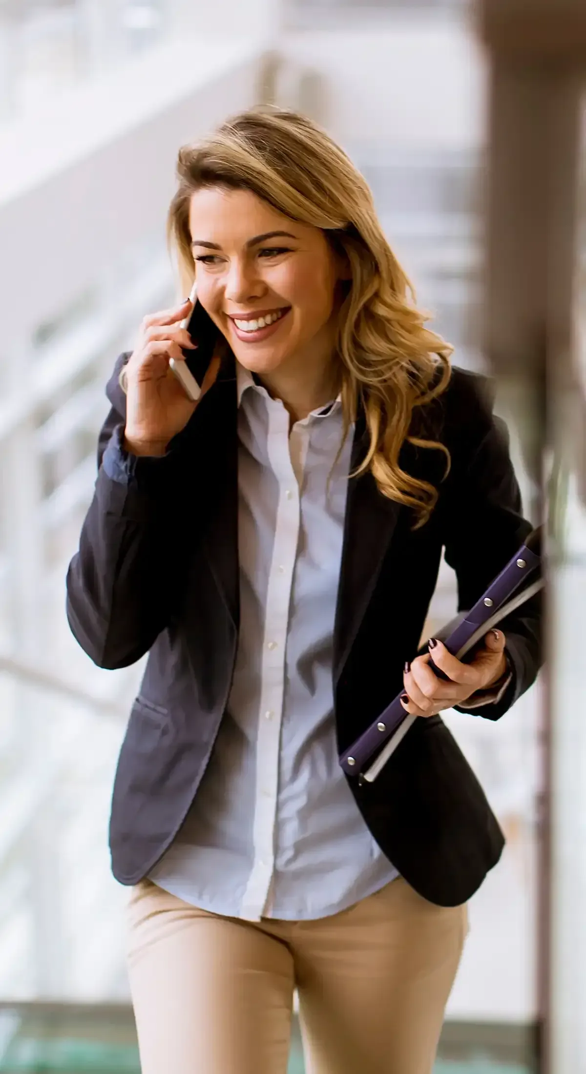 A professional smiling while talking on a phone, walking through an office setting and holding a binder.