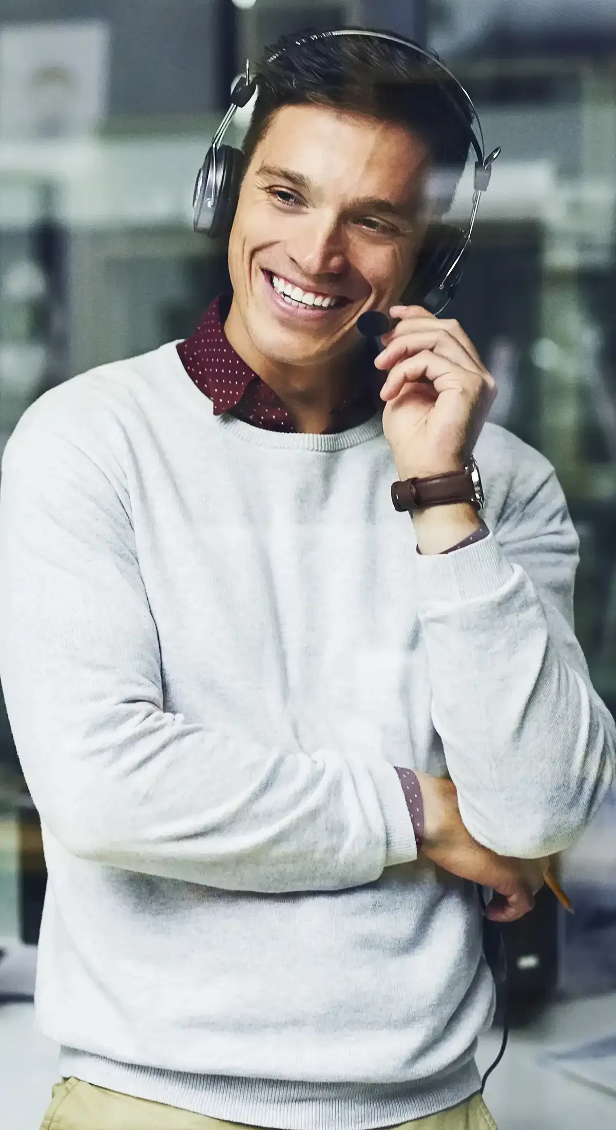A smiling person wearing a headset and a white sweater with a dark collared shirt underneath, standing in an office.