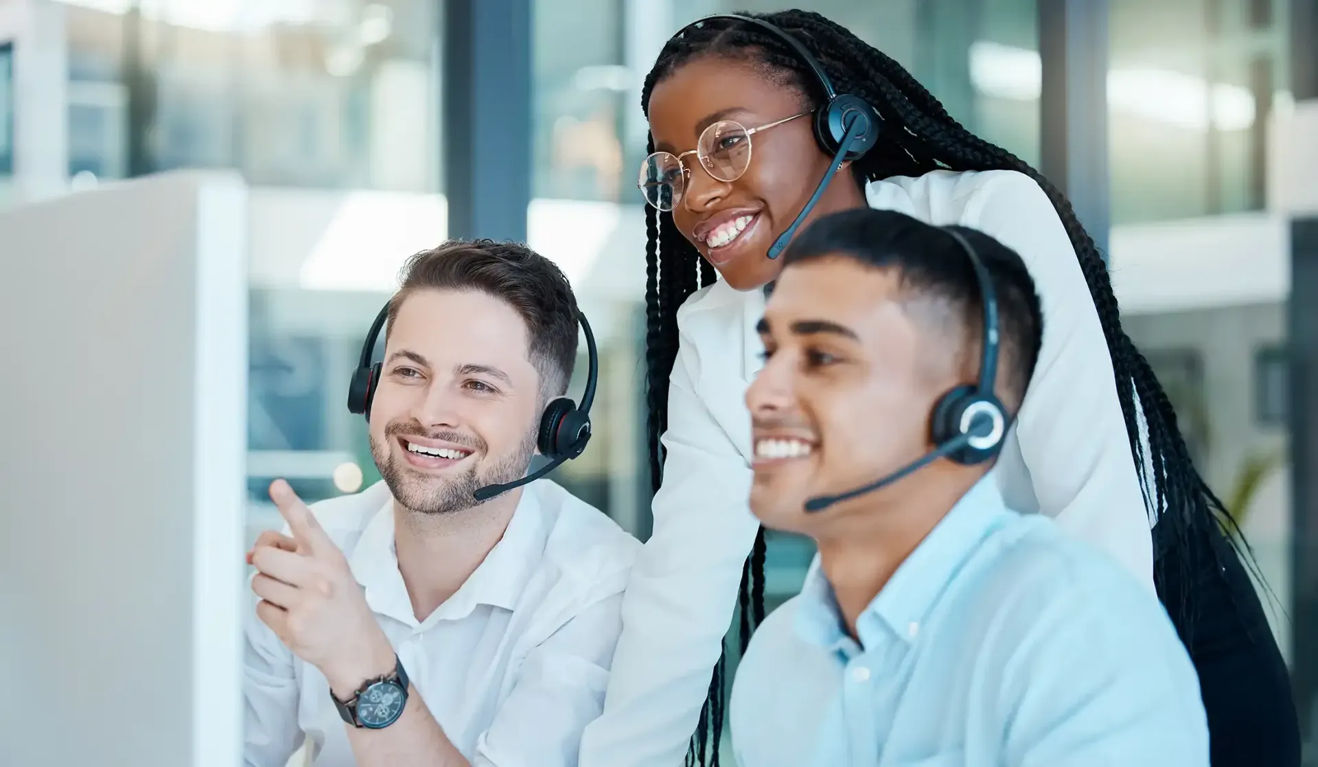 Three colleagues wearing headsets, smiling while pointing at and discussing information on a computer screen.