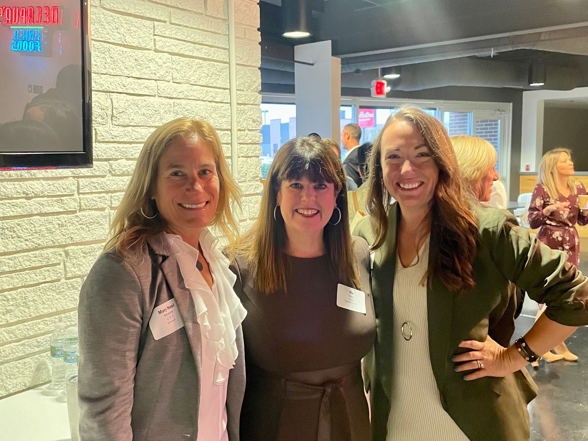 Three women smiling together in a room with white brick walls and bright interior lighting.