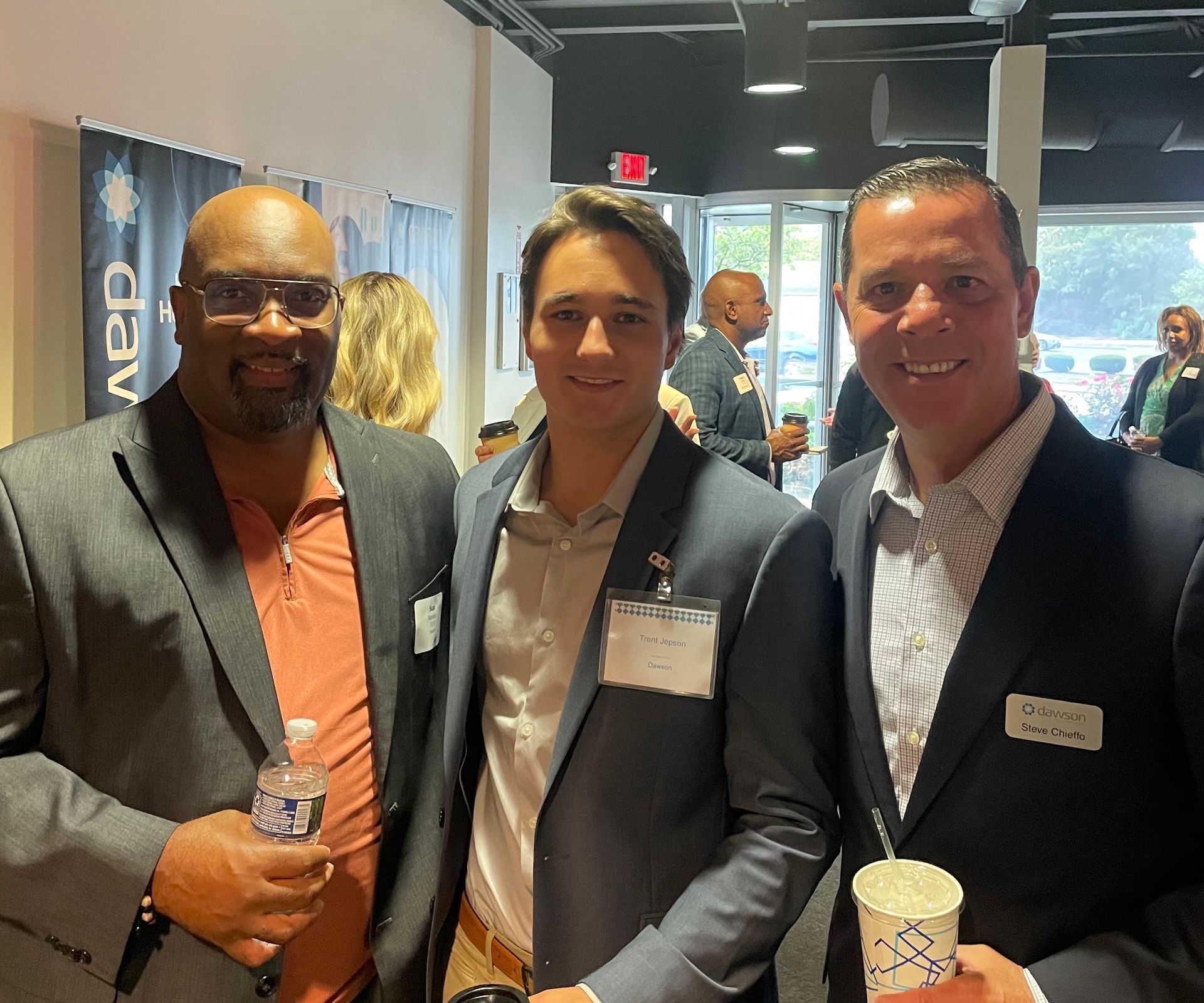 Three people in business casual attire smile at a professional networking event in a brightly lit indoor space.