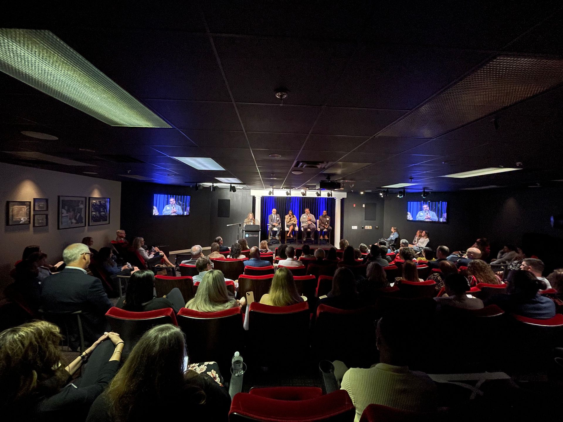 A group of people sits on a lit stage in a dark theater, facing an audience seated in rows of red chairs.