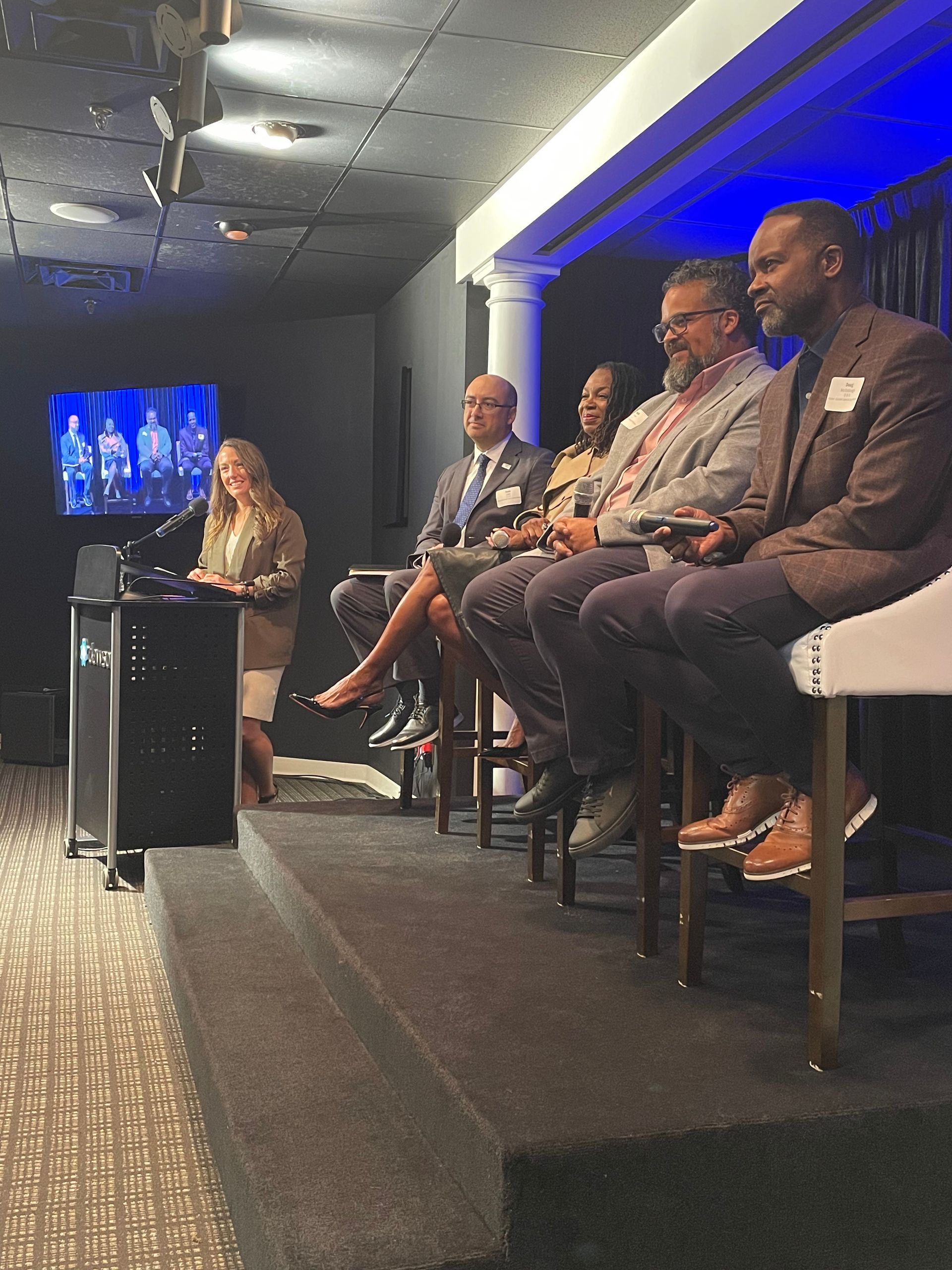 A person stands at a podium addressing an audience while four people sit in a row on a stage during an indoor panel event.