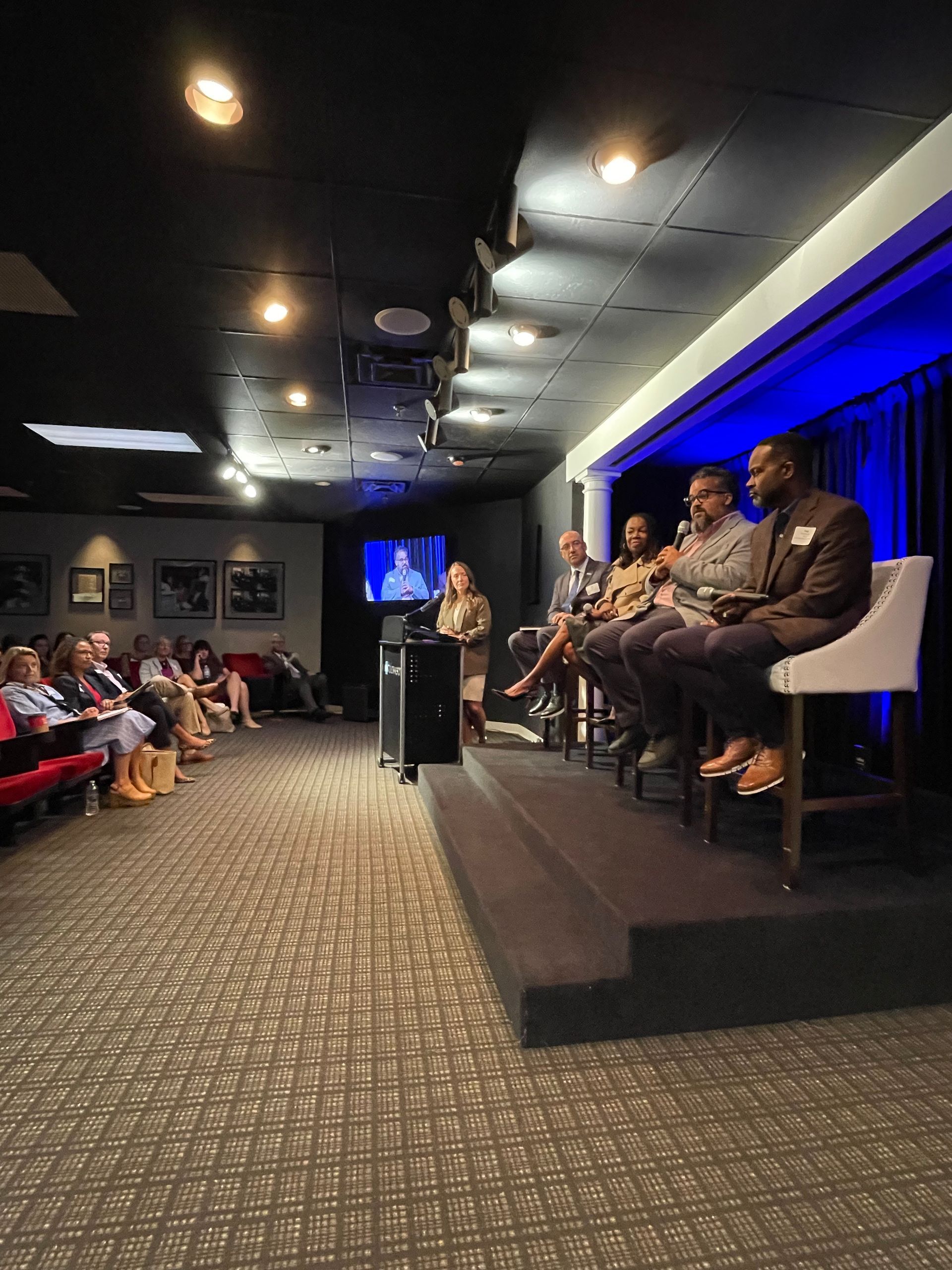A panel of four people sits on a raised stage, speaking to an audience seated in a dimly lit, carpeted meeting room.