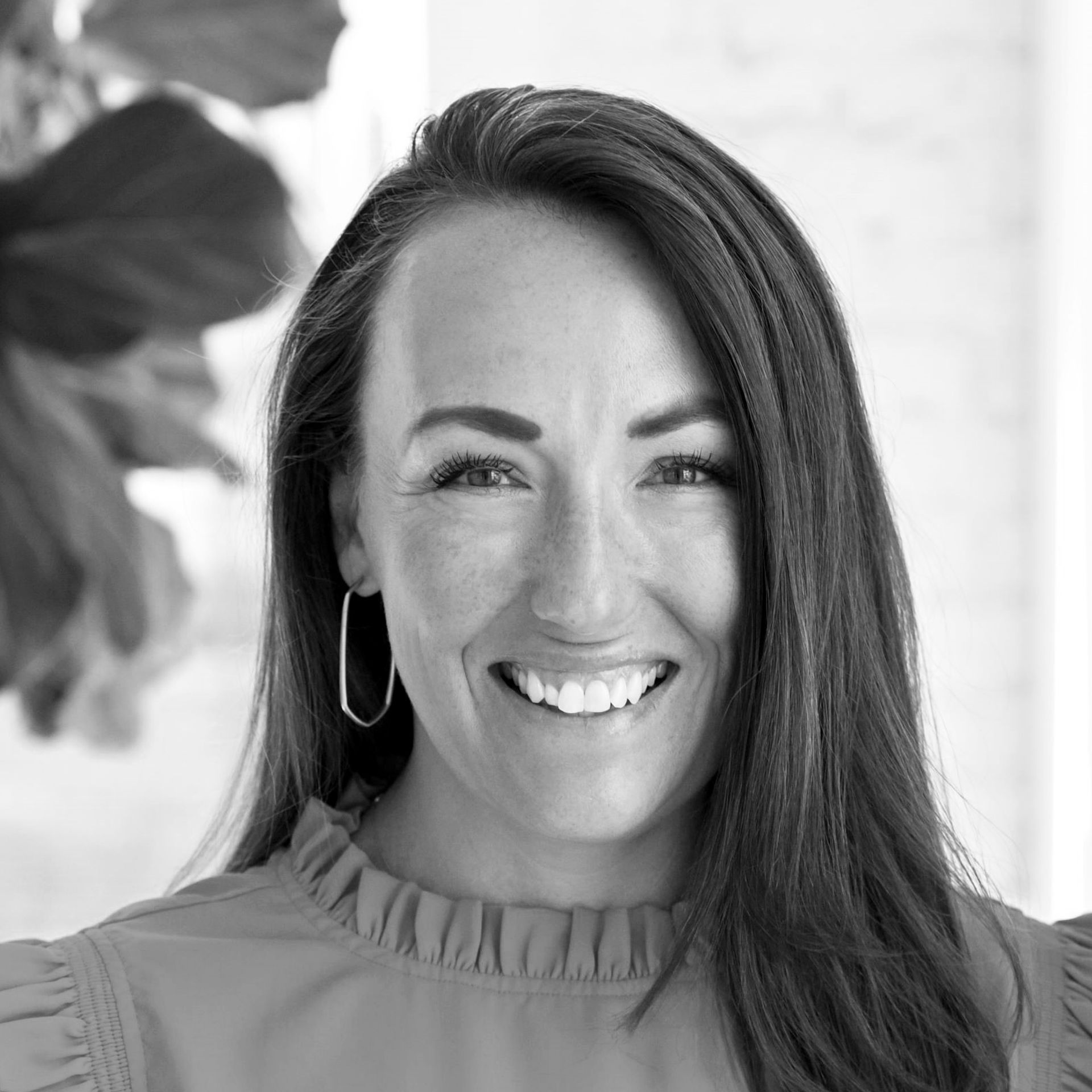 A smiling person with long dark hair, wearing large oval-shaped earrings and a ruffled top against a blurred background.
