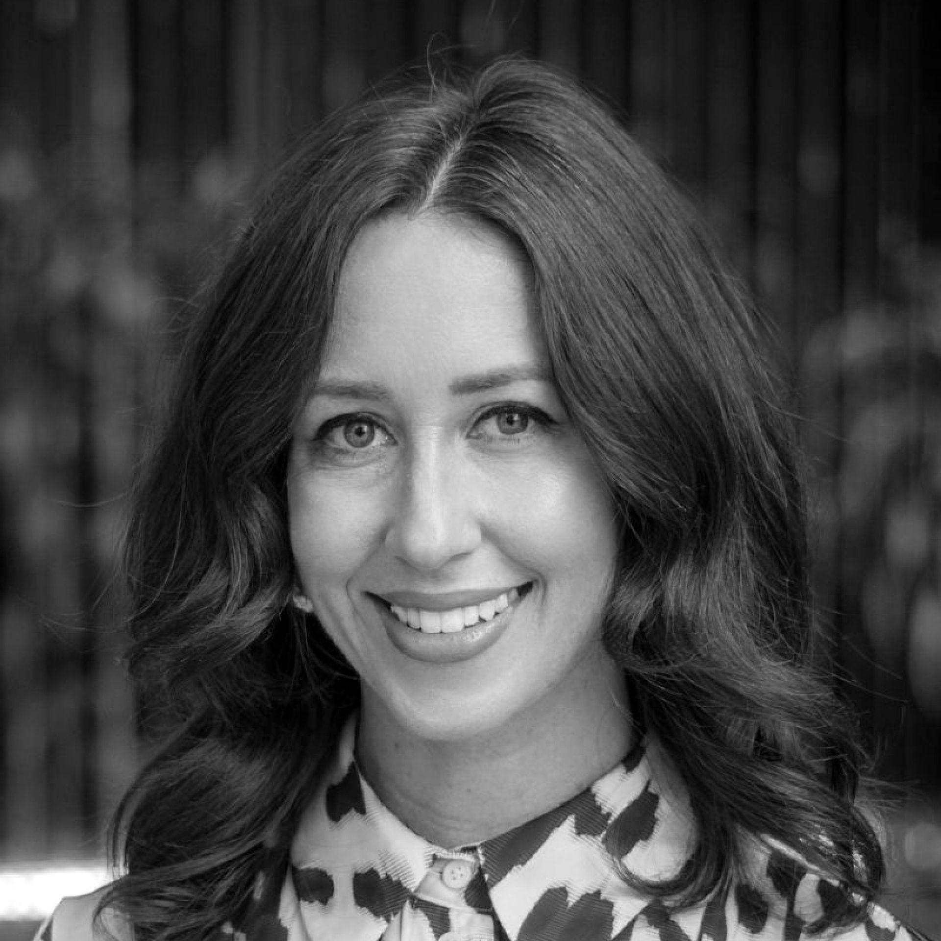 A black-and-white portrait of a person with long wavy hair, smiling and wearing a patterned collared shirt.