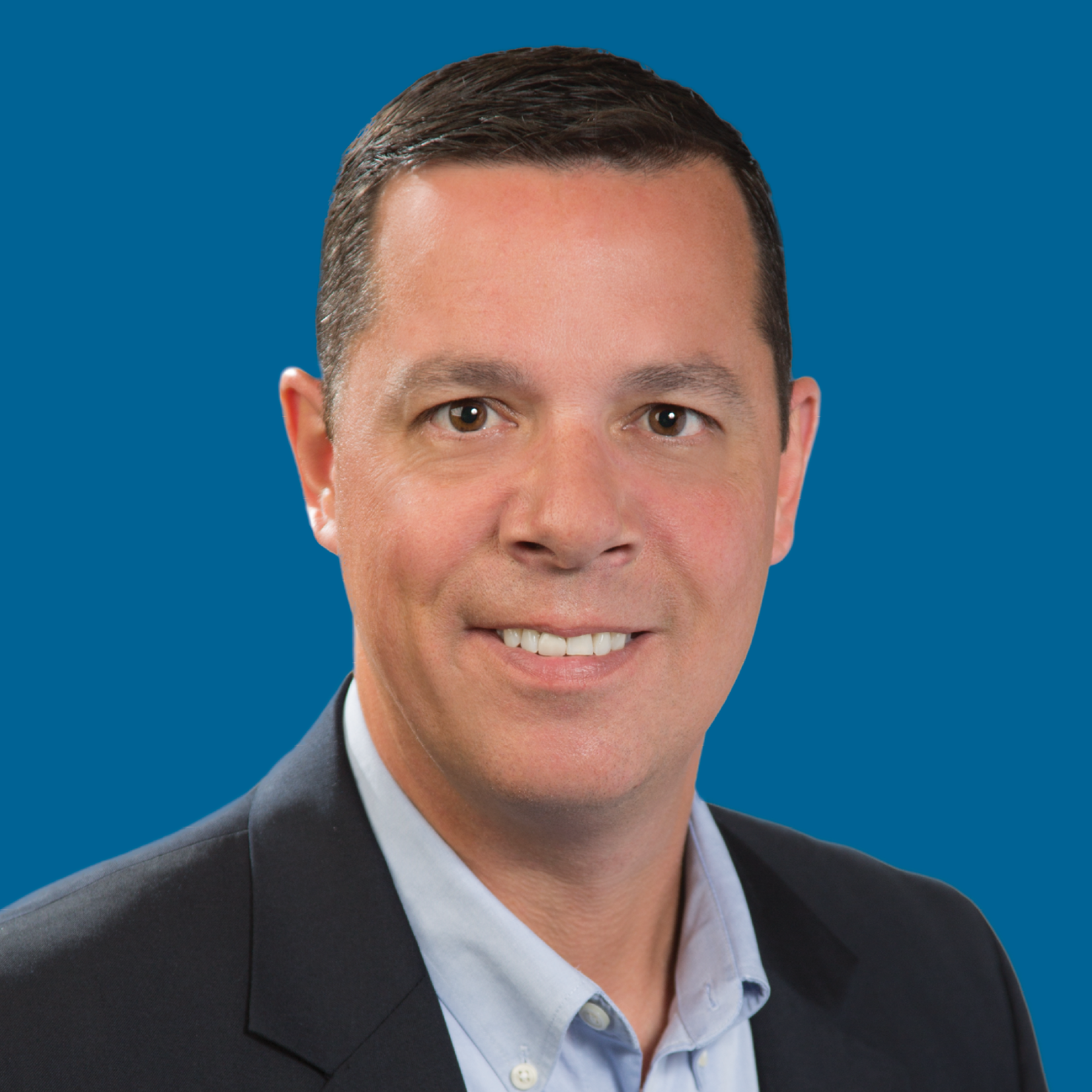A professional headshot of a smiling person wearing a light blue dress shirt and a dark navy suit against a blue background.