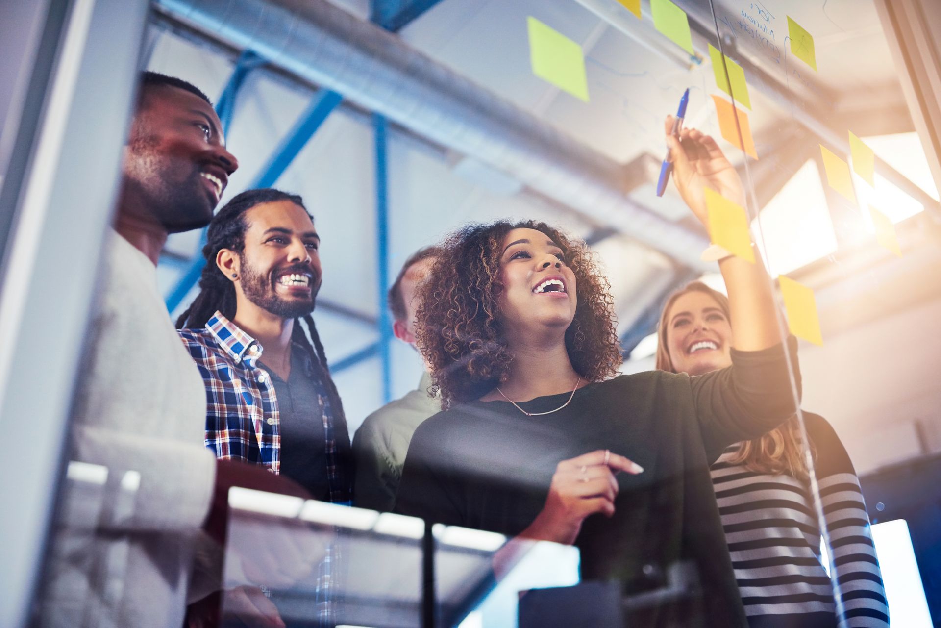 A diverse group of colleagues smiles as they brainstorm by placing sticky notes on a glass office wall.