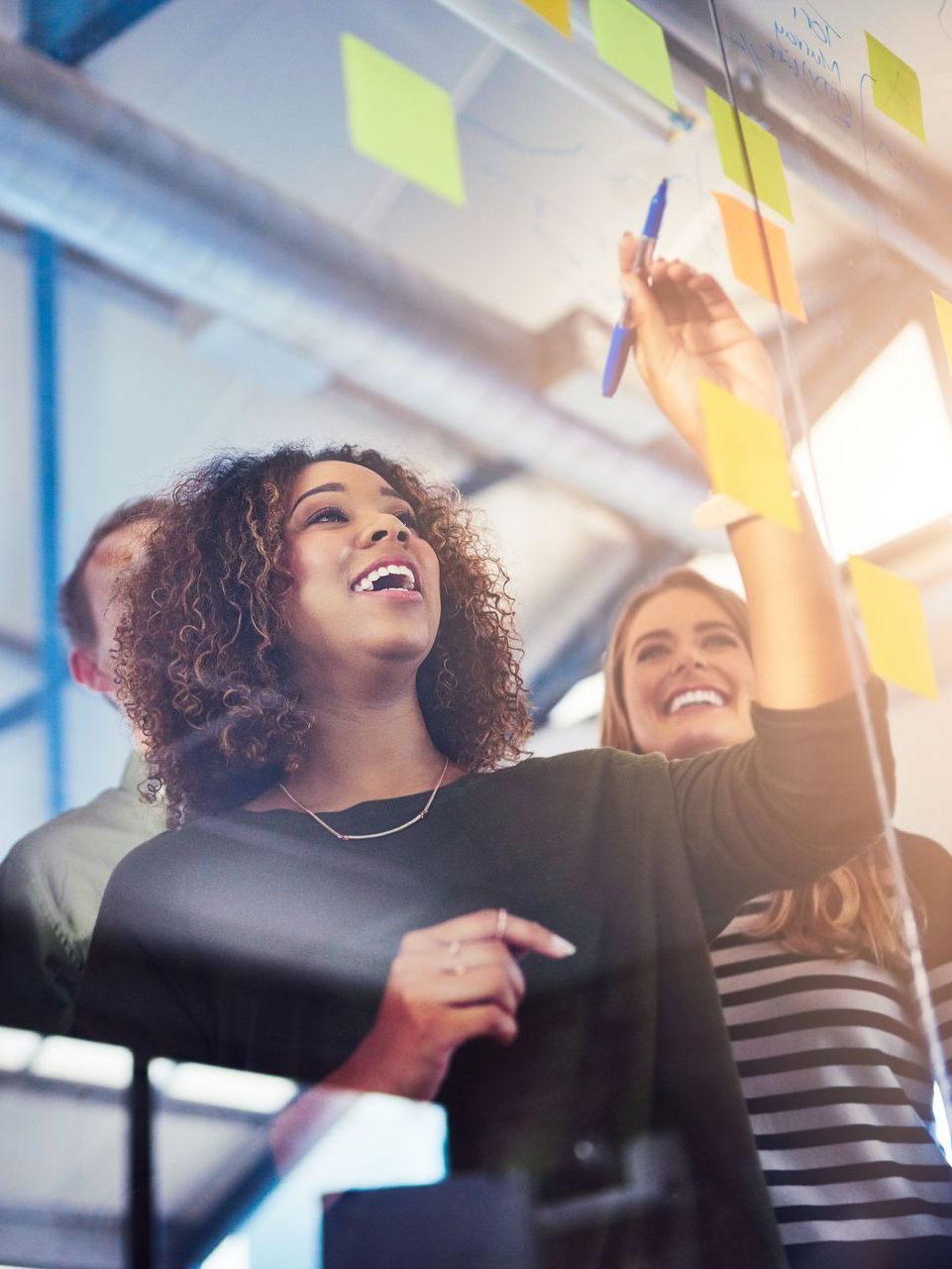 A smiling professional writes on a glass board covered in yellow sticky notes during a collaborative office brainstorming.