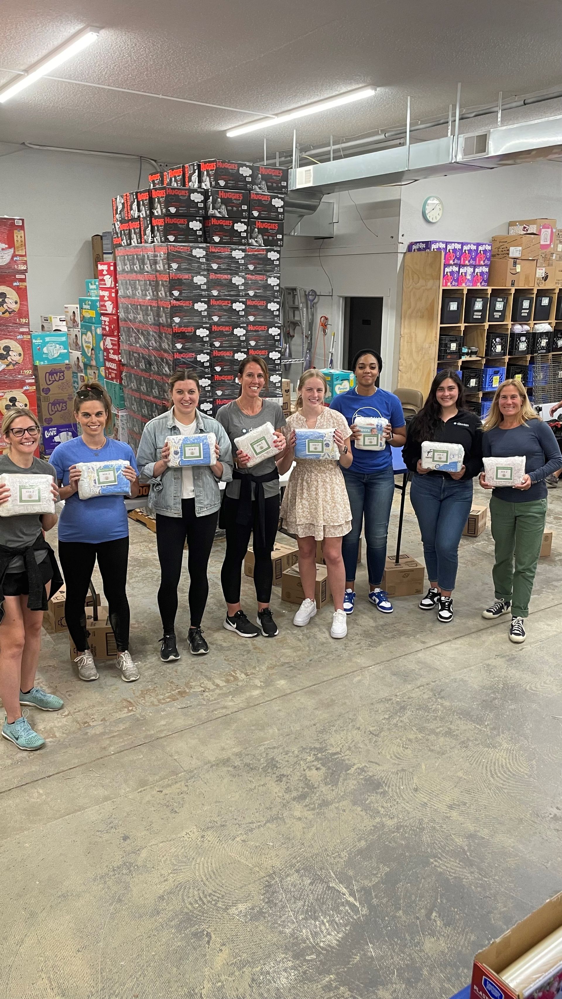 Eight volunteers holding packages stand together in a warehouse filled with stacks of supplies.