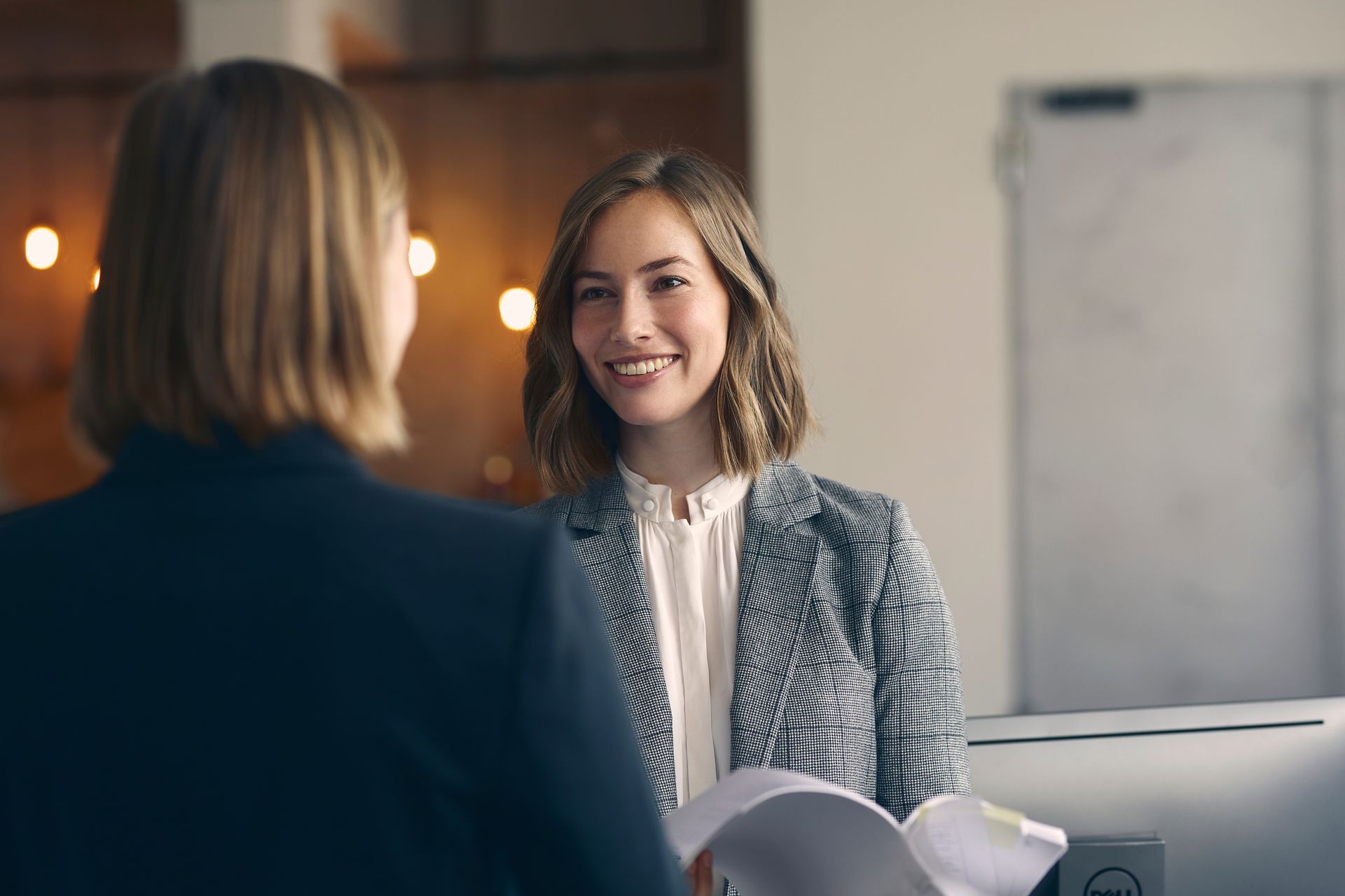 A person in a patterned blazer smiles while conversing with a colleague in a blurred office setting.