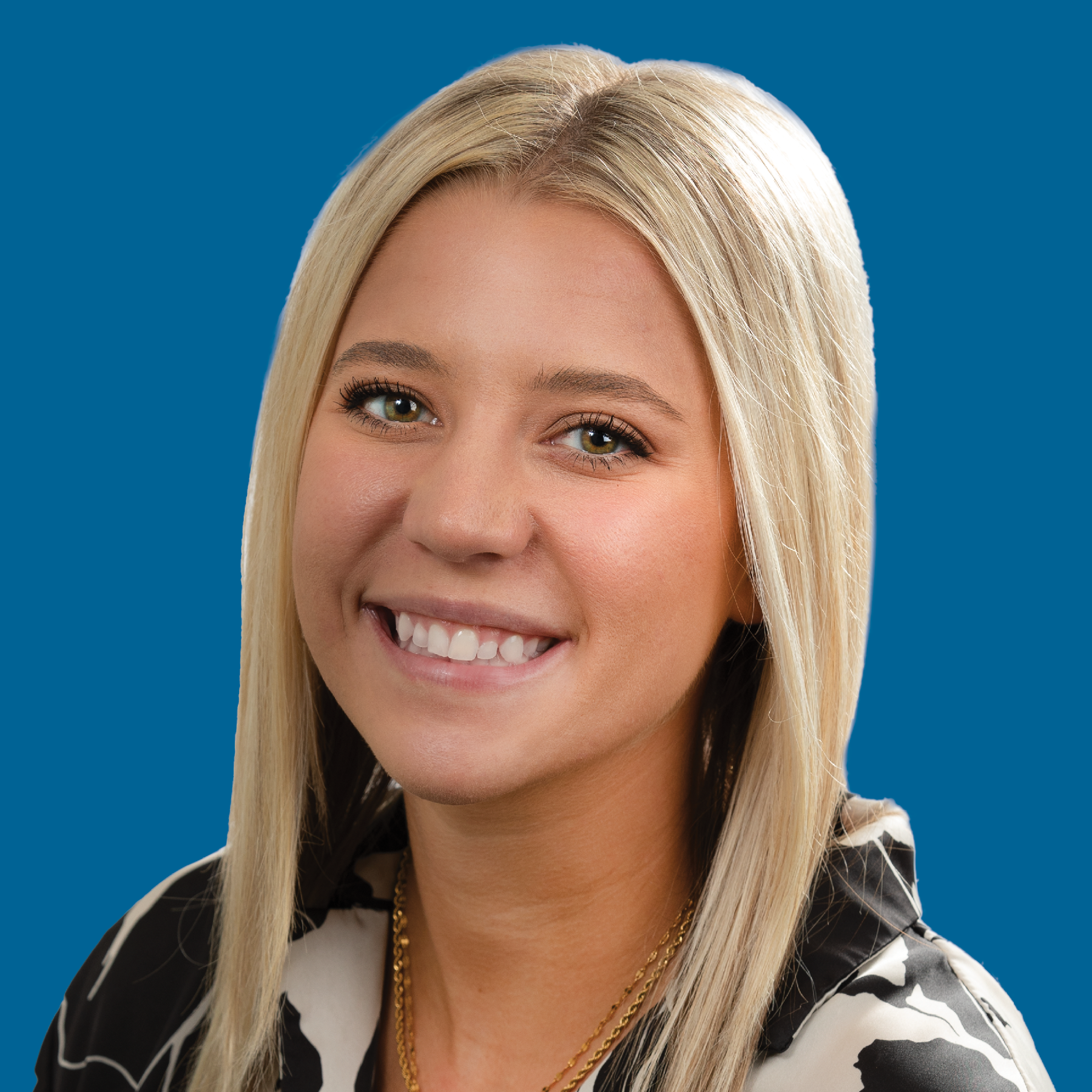 A smiling woman with blonde hair, wearing a patterned top and gold necklace against a solid blue background.