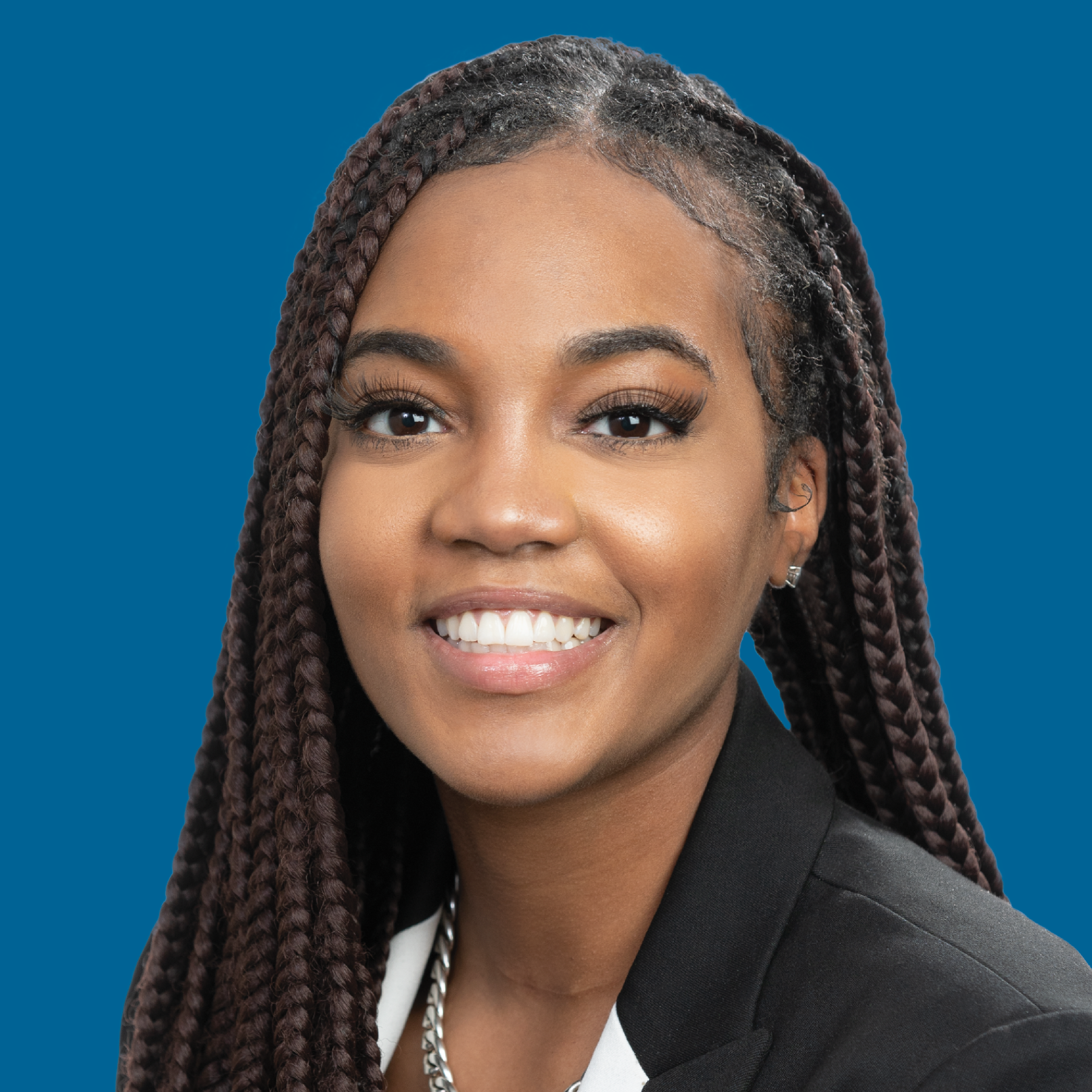 A person with braided hair smiling, wearing a black blazer and white collared shirt against a blue background.