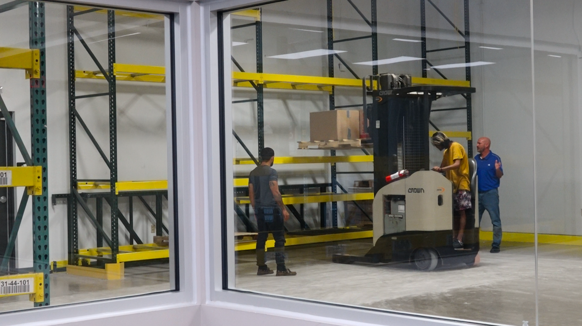 Three people in a warehouse area with yellow shelving as one operates a forklift to lift a pallet of boxes.