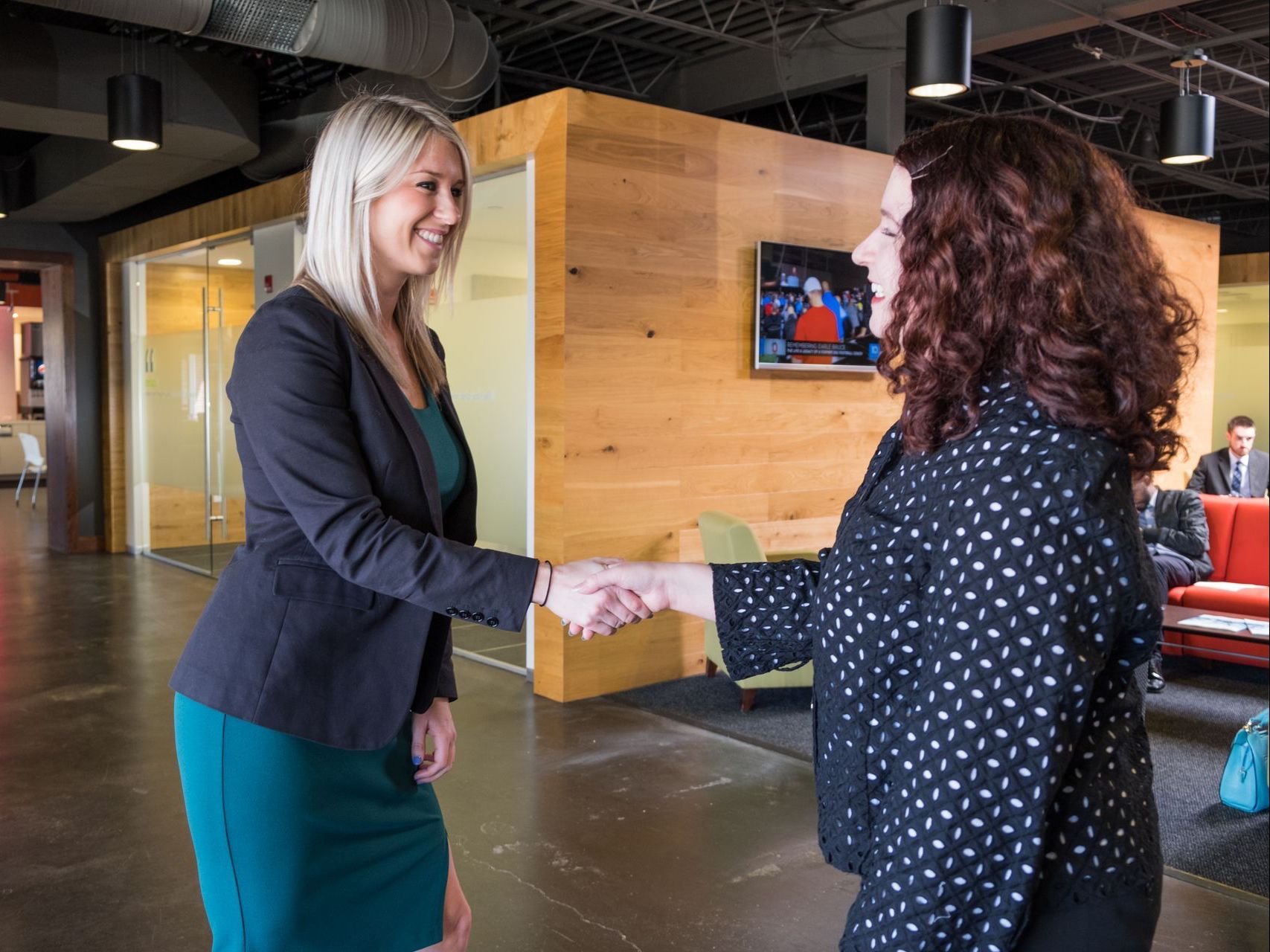 Two professionals in business attire shake hands in a modern, open-plan office setting with wooden wall accents.