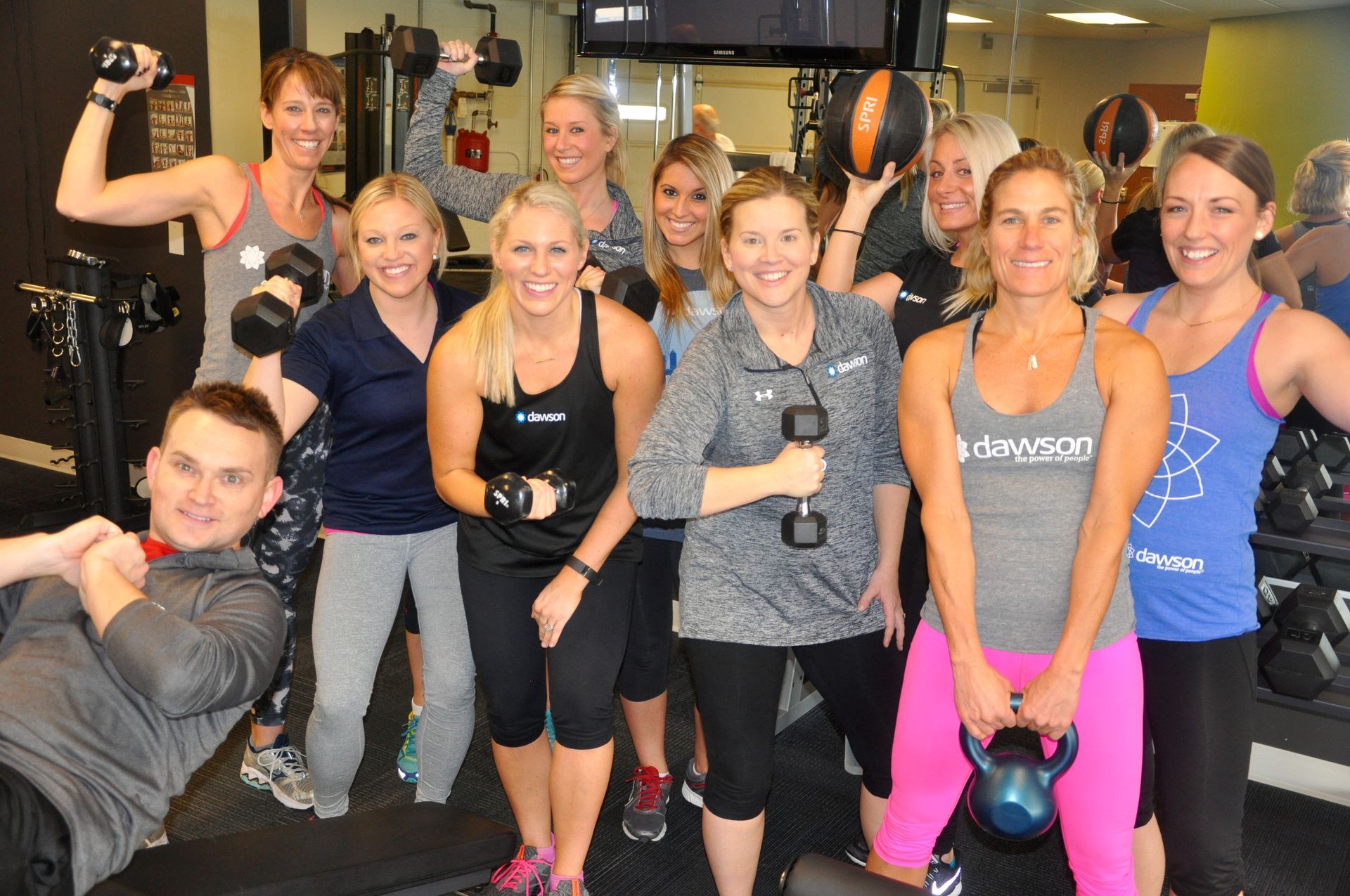 A diverse group of smiling people in a gym, posing with various fitness equipment while dressed in workout attire.