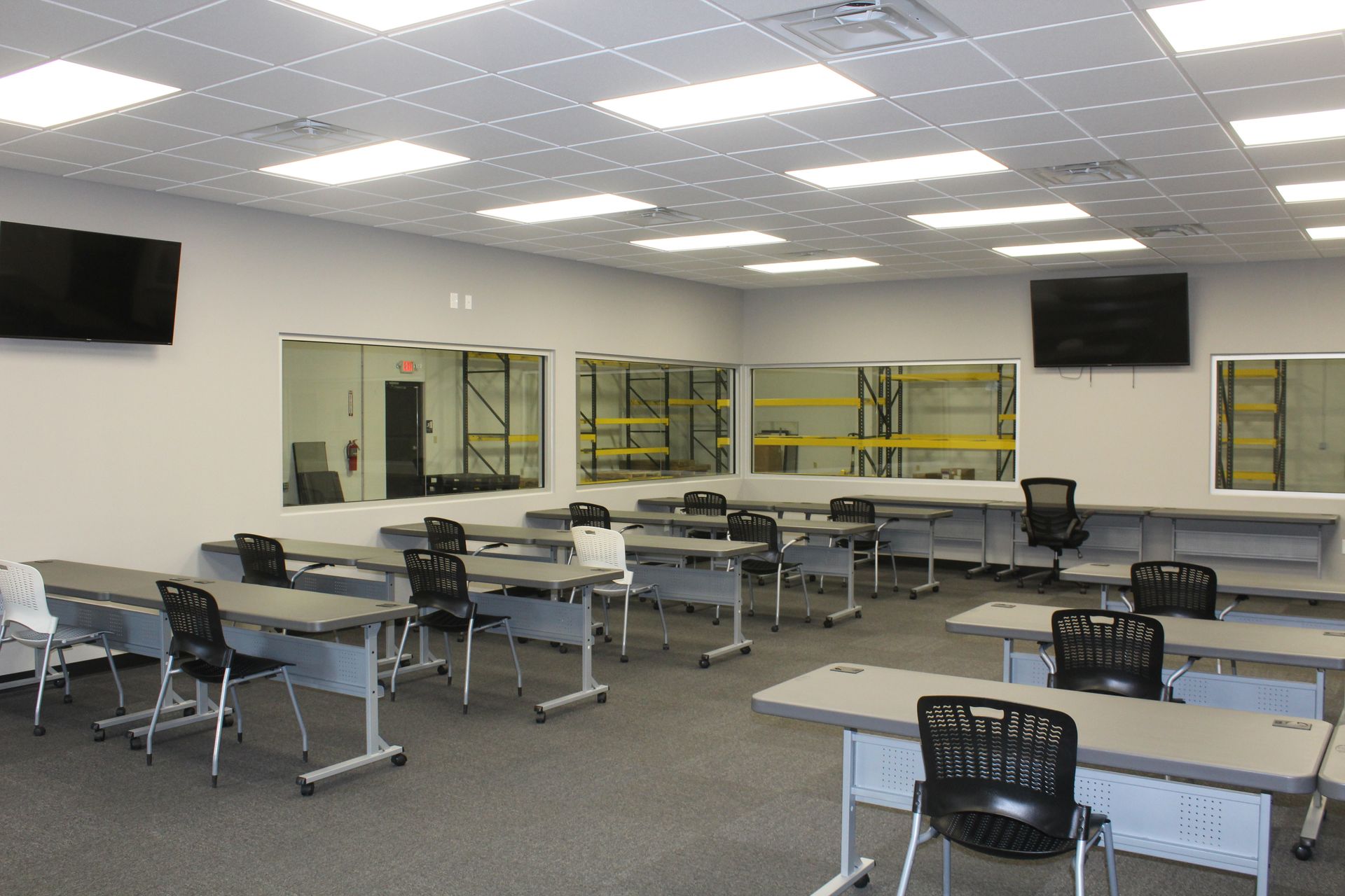 A sterile, brightly lit training room with rows of grey tables and black chairs facing windows looking into a warehouse.