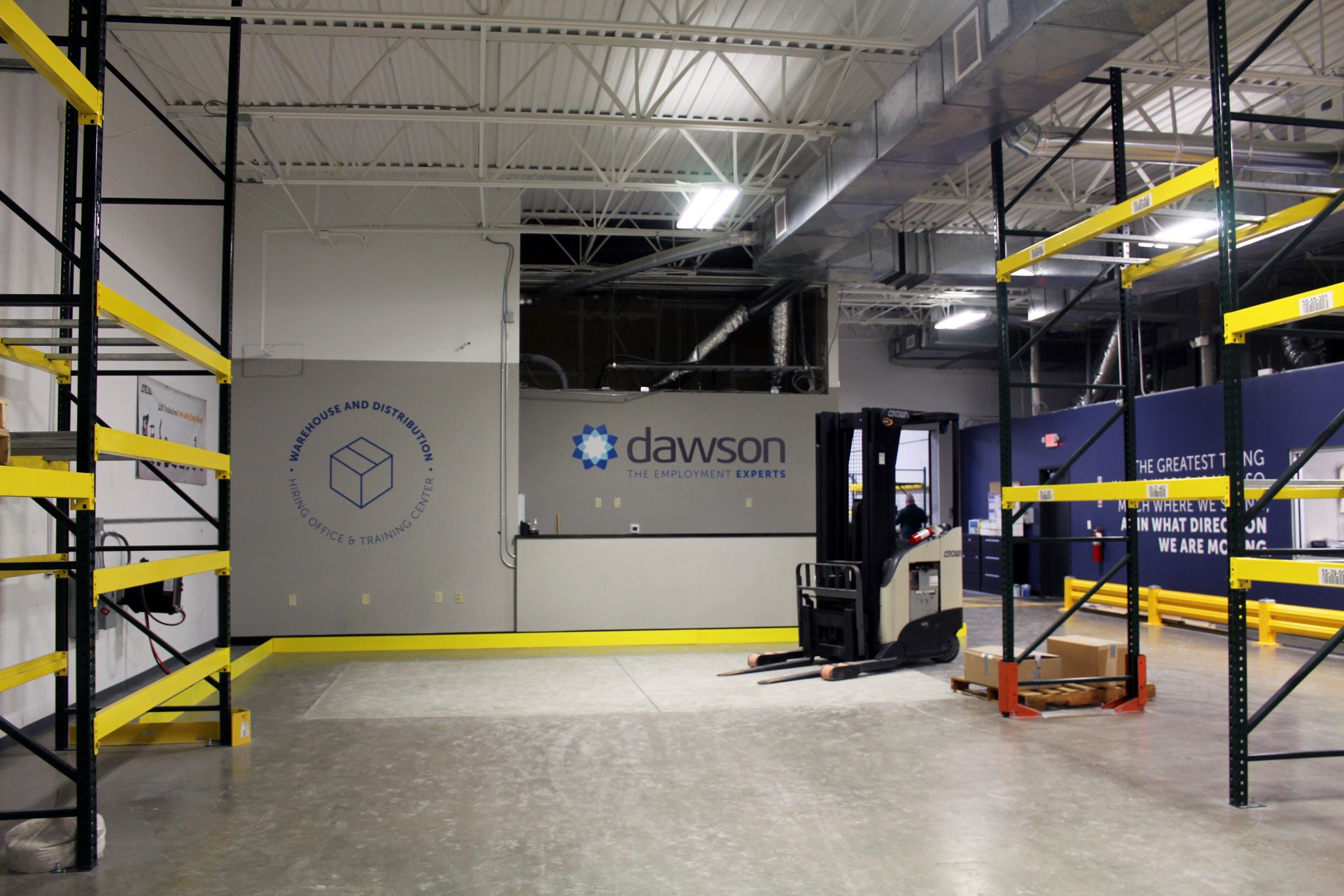 A forklift sits in a warehouse between rows of yellow and black metal storage shelving, with a Dawson logo on the wall.