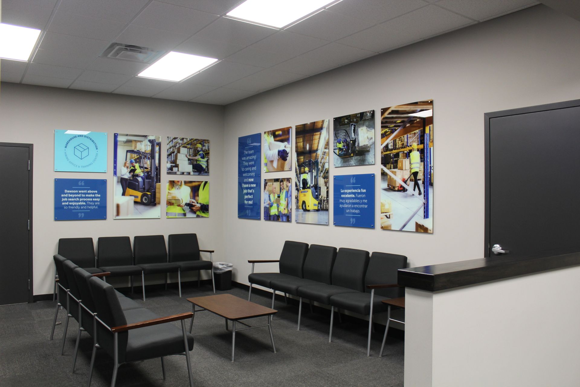 A waiting area with black chairs arranged around a coffee table, featuring wall art depicting warehouse operations.