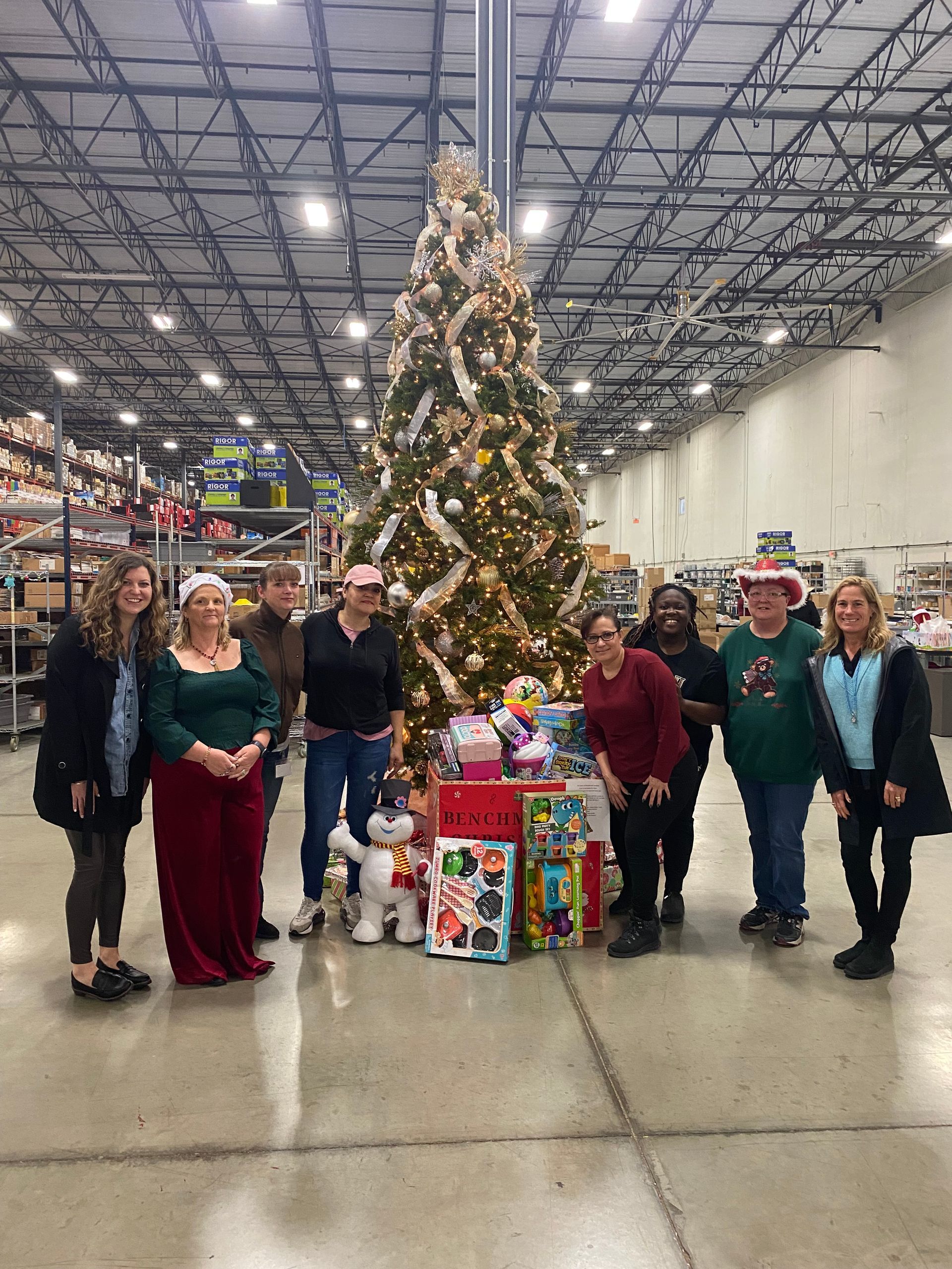 A group of eight people stand together in a warehouse around a tall, decorated Christmas tree with gifts at its base.
