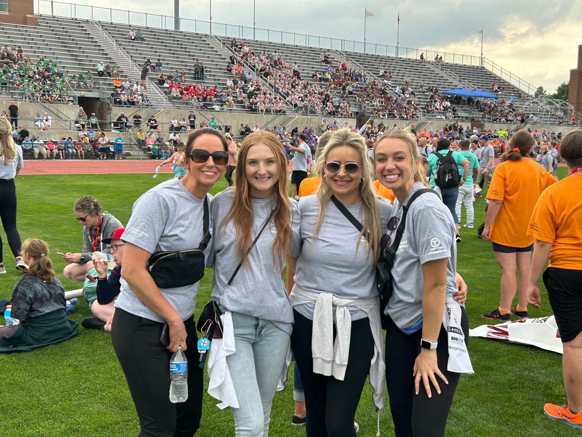 Four people in matching gray t-shirts stand smiling on a grassy field in front of a stadium filled with spectators.