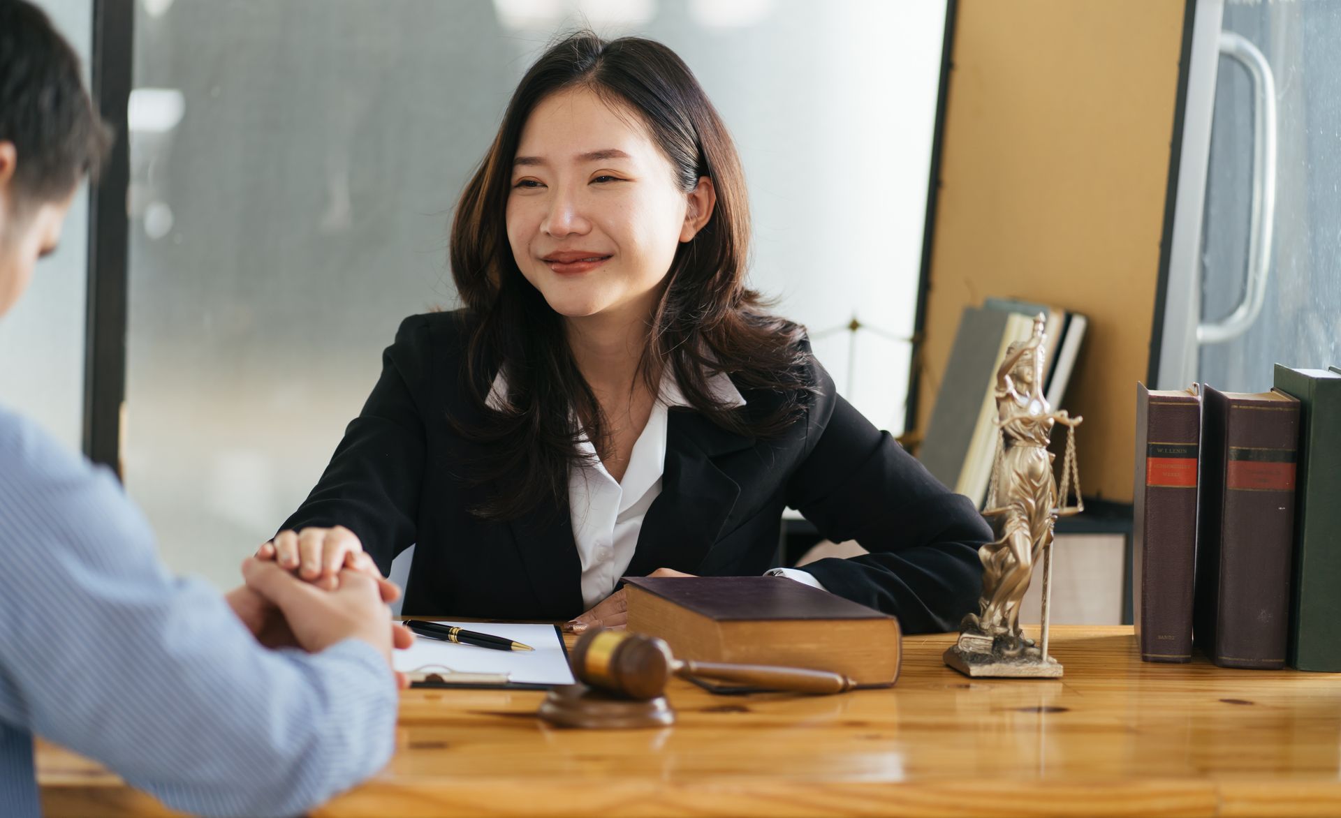 A legal professional sits at a desk with a gavel and scales of justice, listening to a client in an office setting.