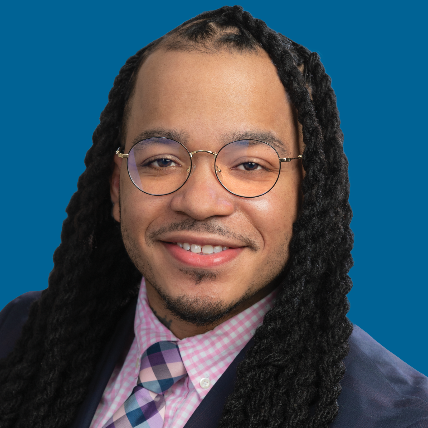 Headshot of a person with long braided hair wearing round glasses, a plaid pink shirt, and a tie against a blue background.