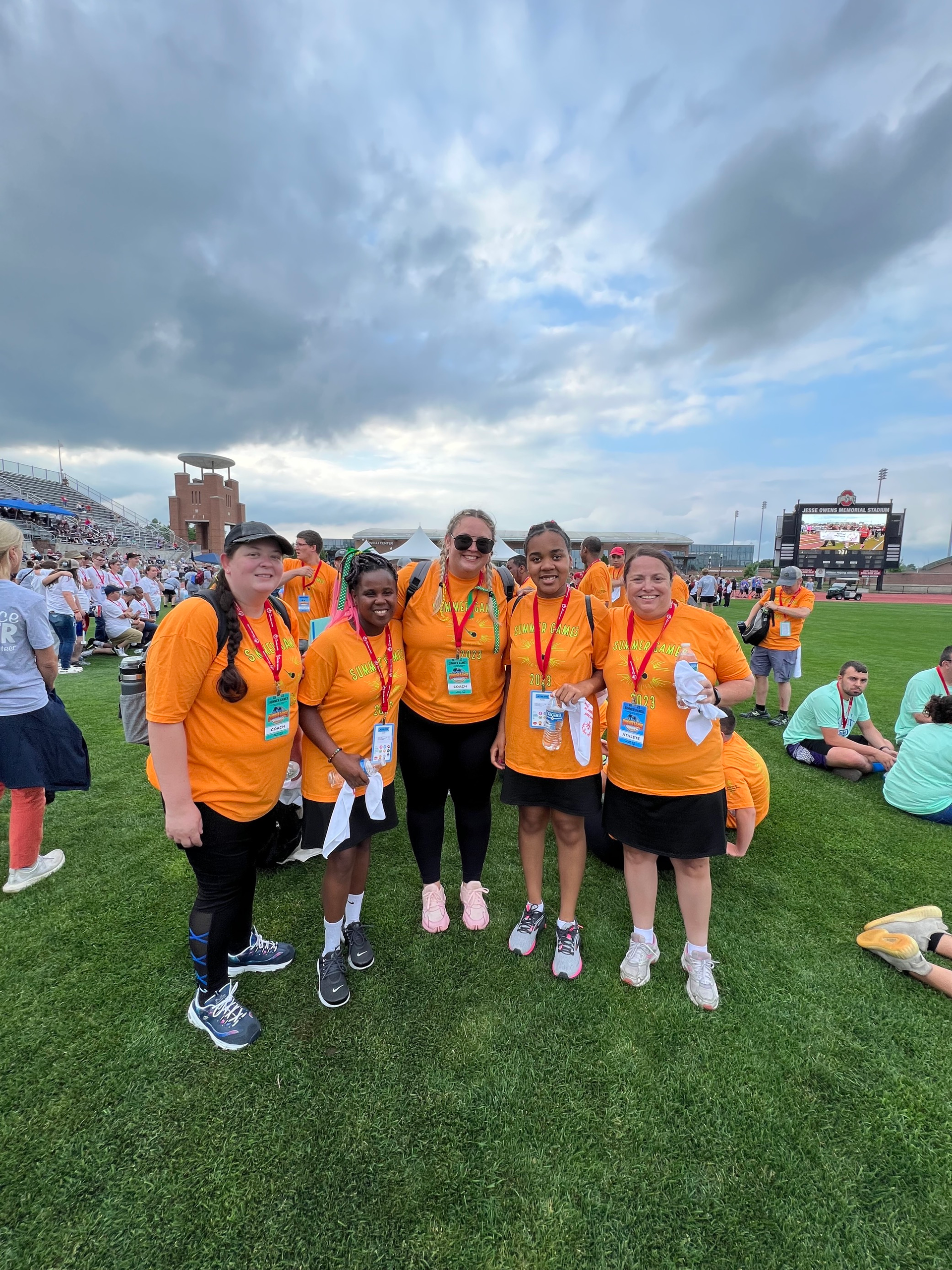 Five people in matching orange t-shirts and lanyards stand together on a grassy field at a stadium under a cloudy sky.