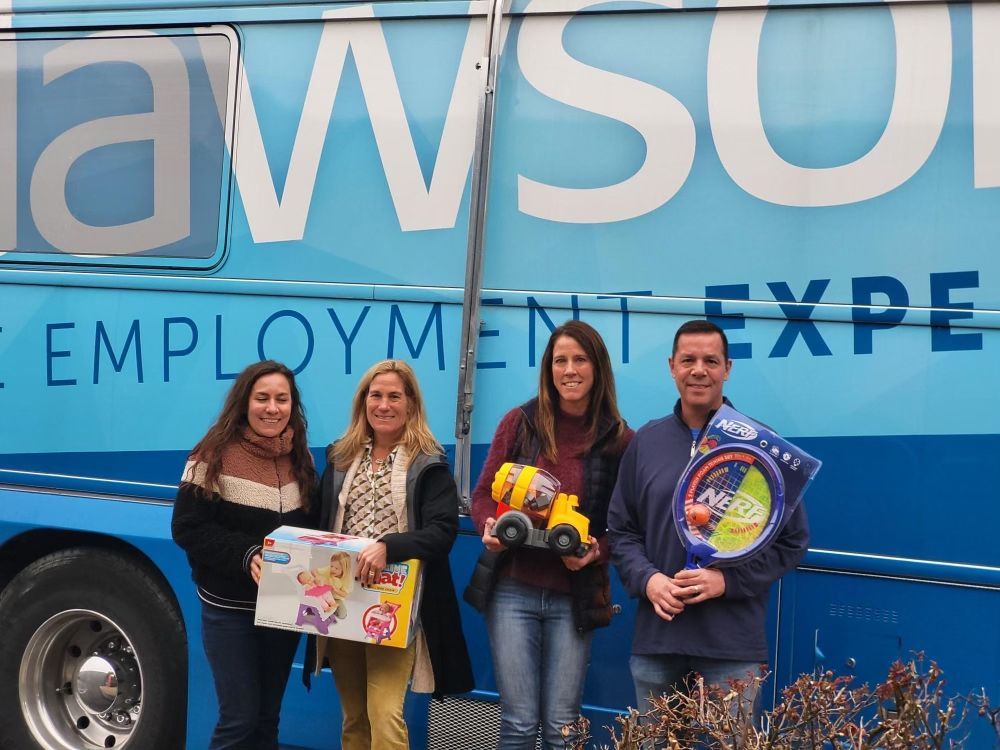 Four people stand in front of a blue Lawson employment bus holding wrapped toys and games for a donation drive.