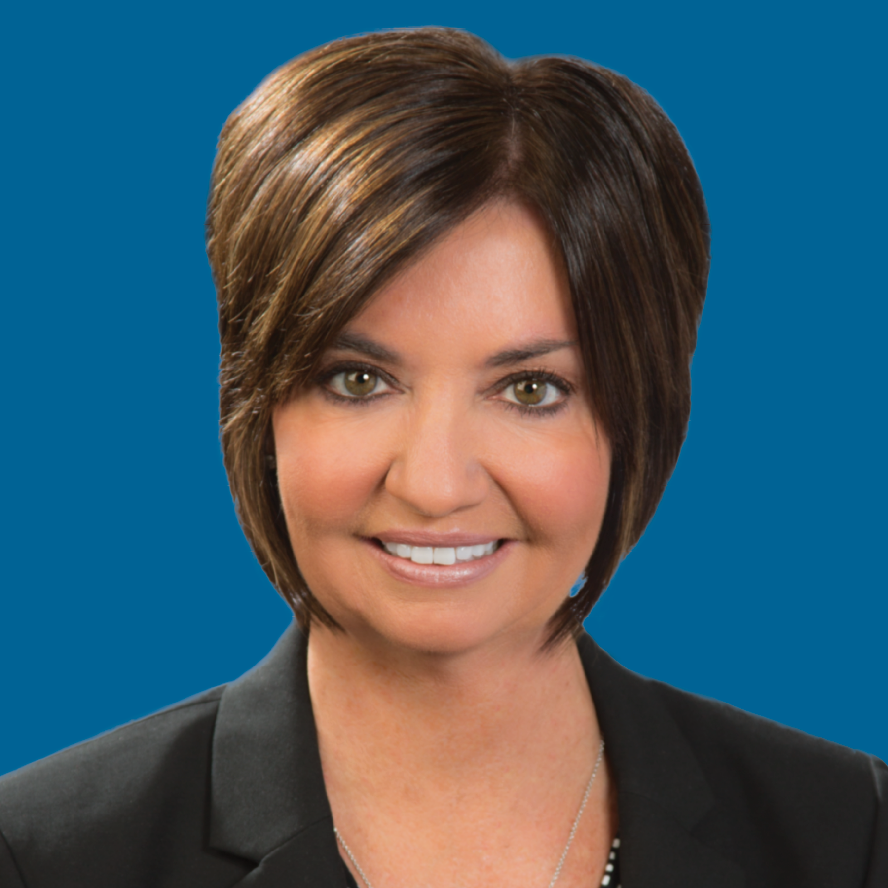 Headshot of a person with shoulder-length brown hair, smiling, wearing a black blazer against a solid blue background.