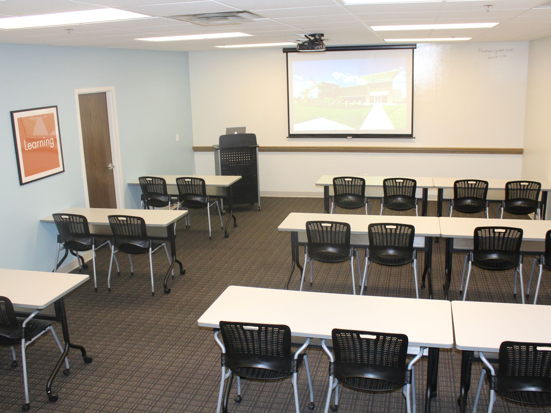 A classroom with rows of tables and black chairs facing a projection screen on a white wall.