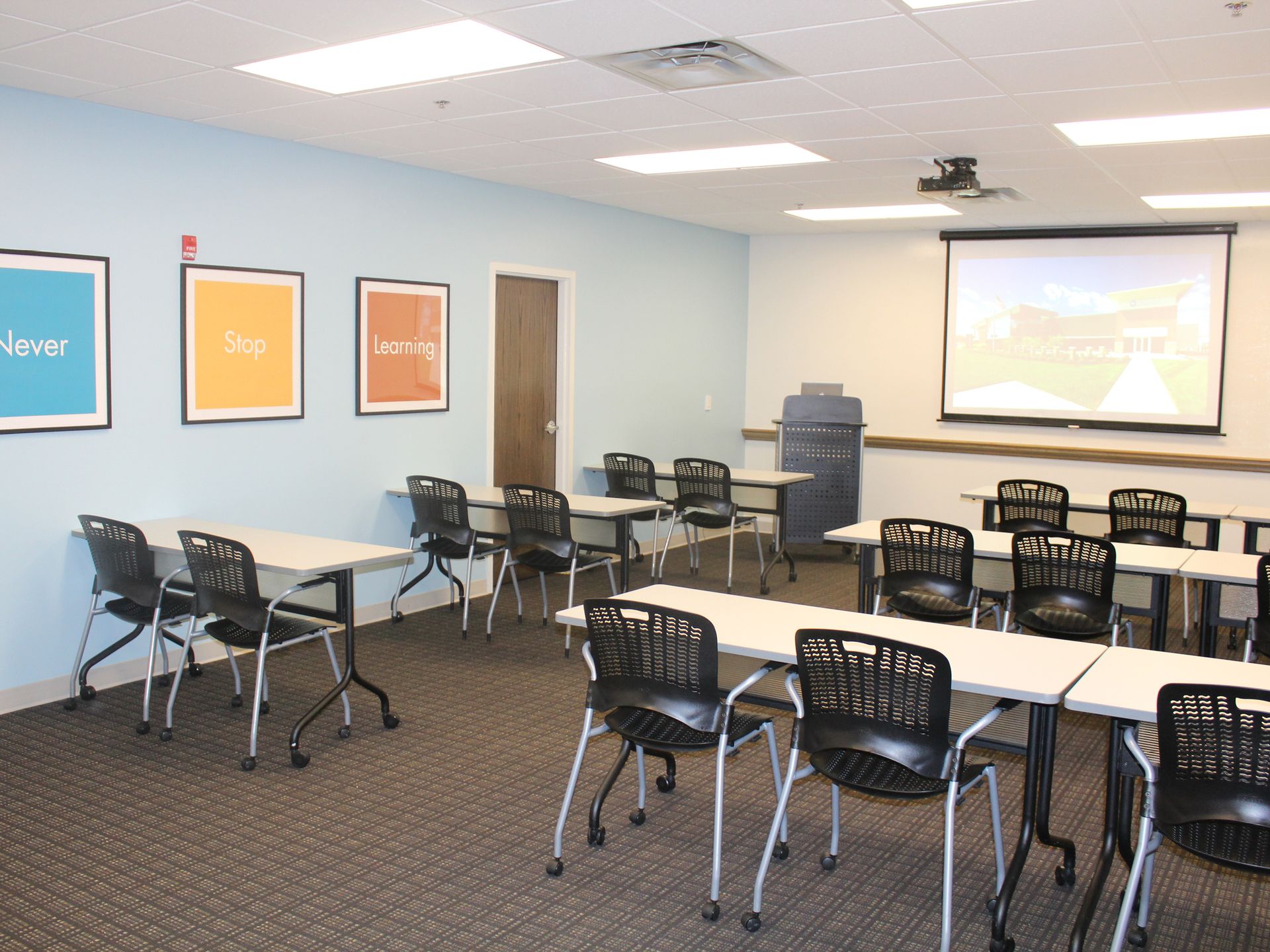 A bright meeting room with rows of tables and chairs, a projector screen, and three colorful framed posters on the wall.